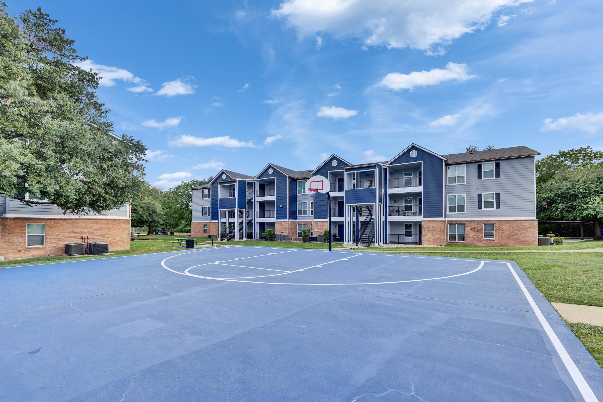 A view of an apartment complex featuring modern blue and beige buildings. In the foreground, there is a blue basketball court with a hoop. The background includes green grass and trees under a partly cloudy sky.