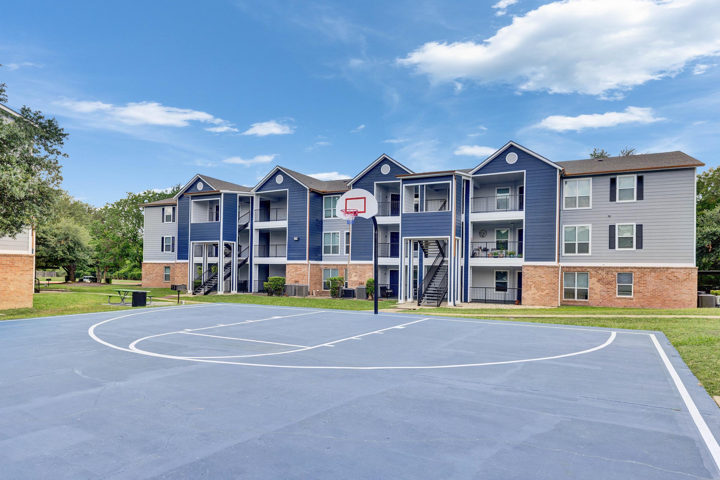 Basketball court in the foreground with a blue surface and a hoop. In the background, there are three-story residential apartment buildings painted in blue and beige, surrounded by green grass and trees under a clear blue sky.