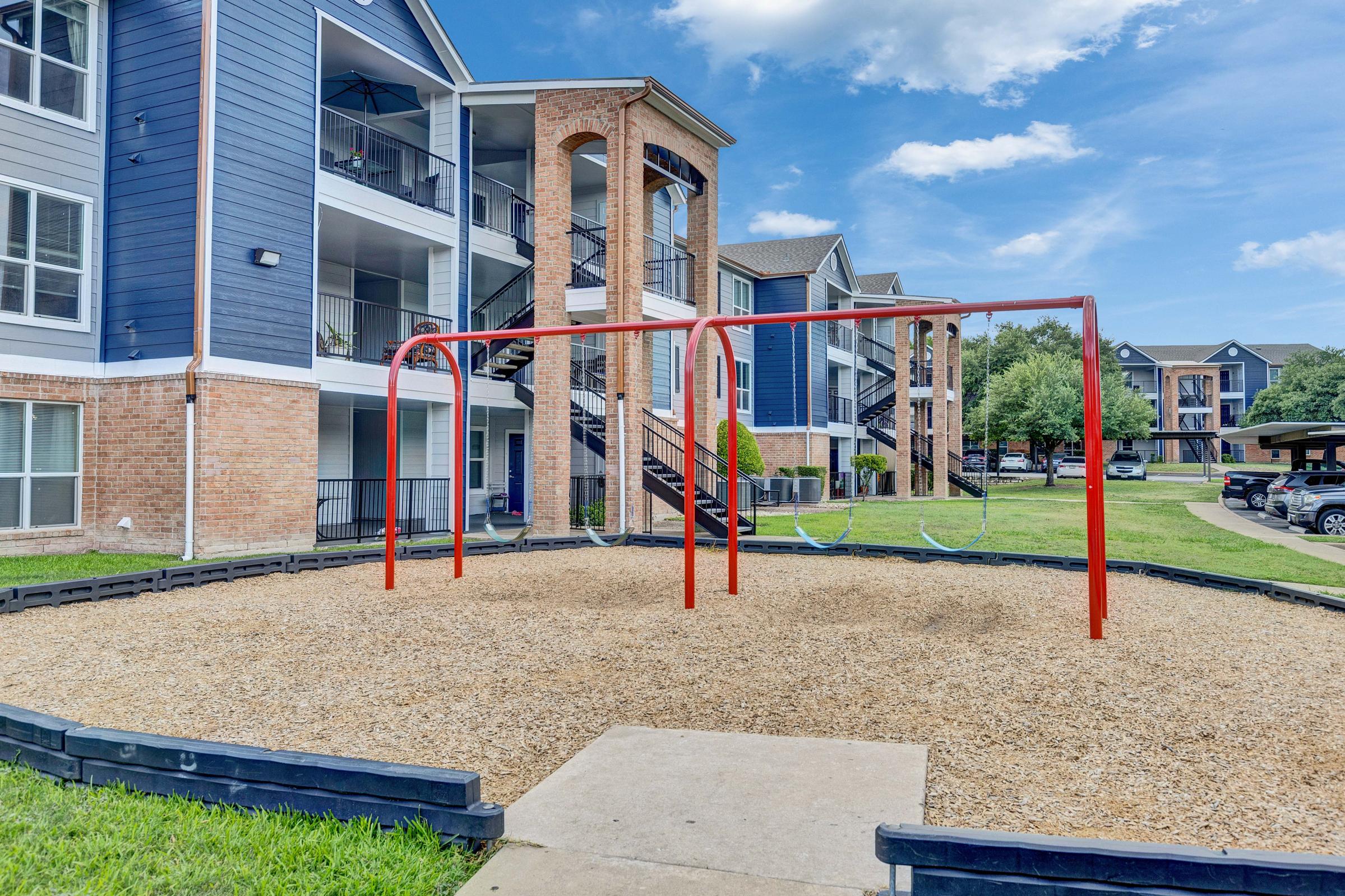 A playground area featuring two red swing sets surrounded by a gravel surface, located in front of a multi-story residential building. Lush green grass and parking spaces are visible in the background under a partly cloudy sky.