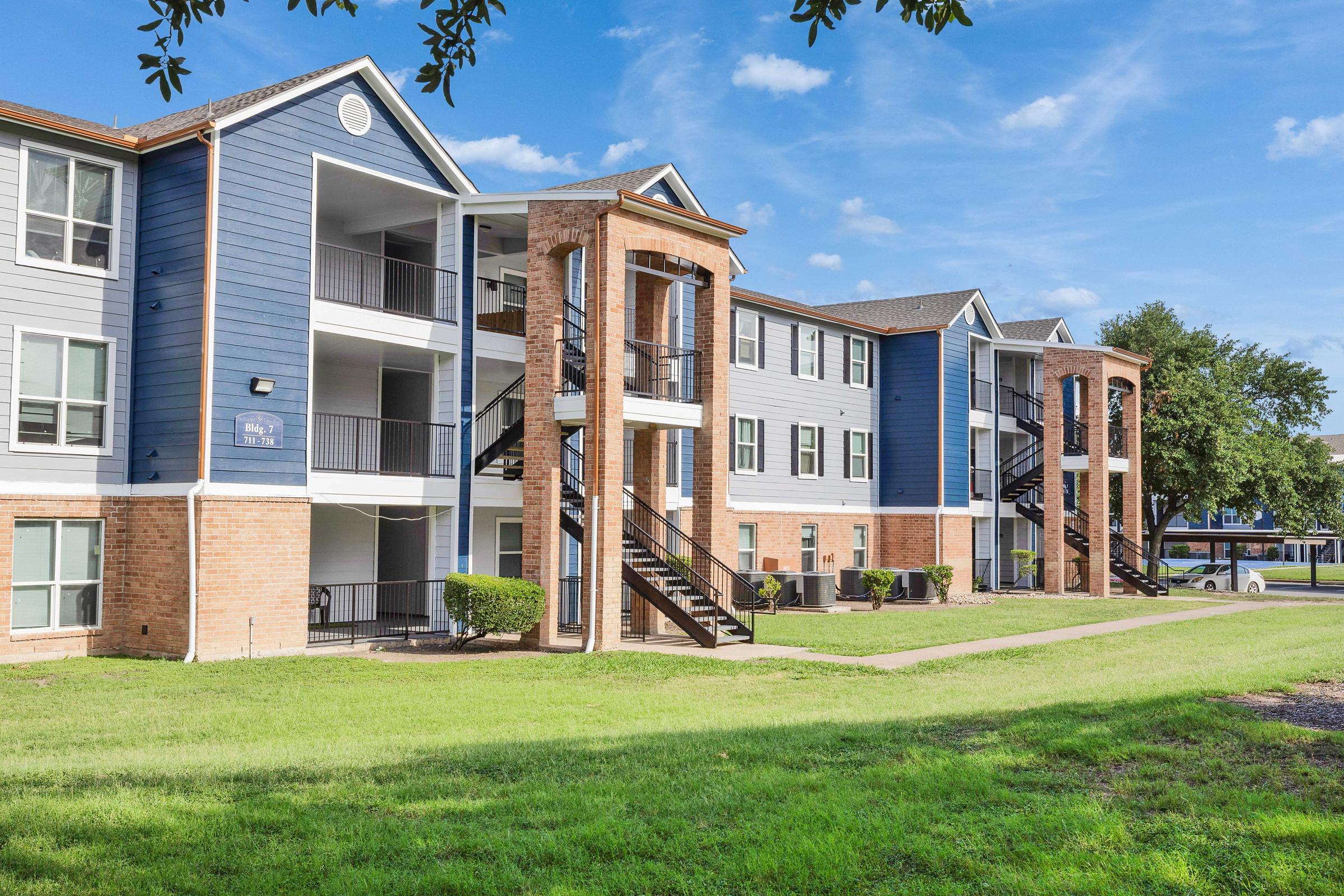 Exterior view of a modern apartment building featuring three floors. The building has a mix of blue and brick facades, with balconies and metal staircases. The surrounding area includes green grass and a clear blue sky, creating a bright and inviting atmosphere.