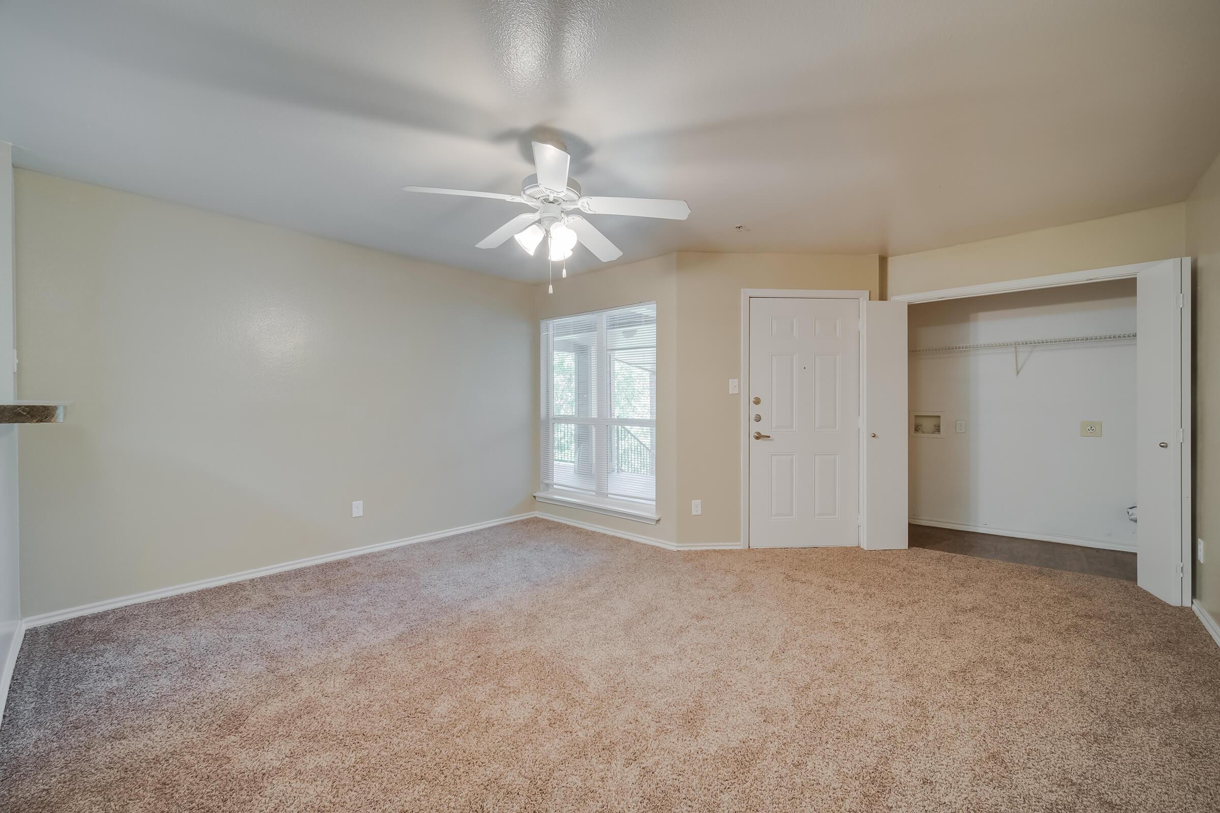 Spacious living area with light beige walls and plush carpet. A ceiling fan hangs from the ceiling, providing ventilation. There is a large window allowing natural light, and a closet space can be seen in the corner. The overall atmosphere is inviting and well-lit.