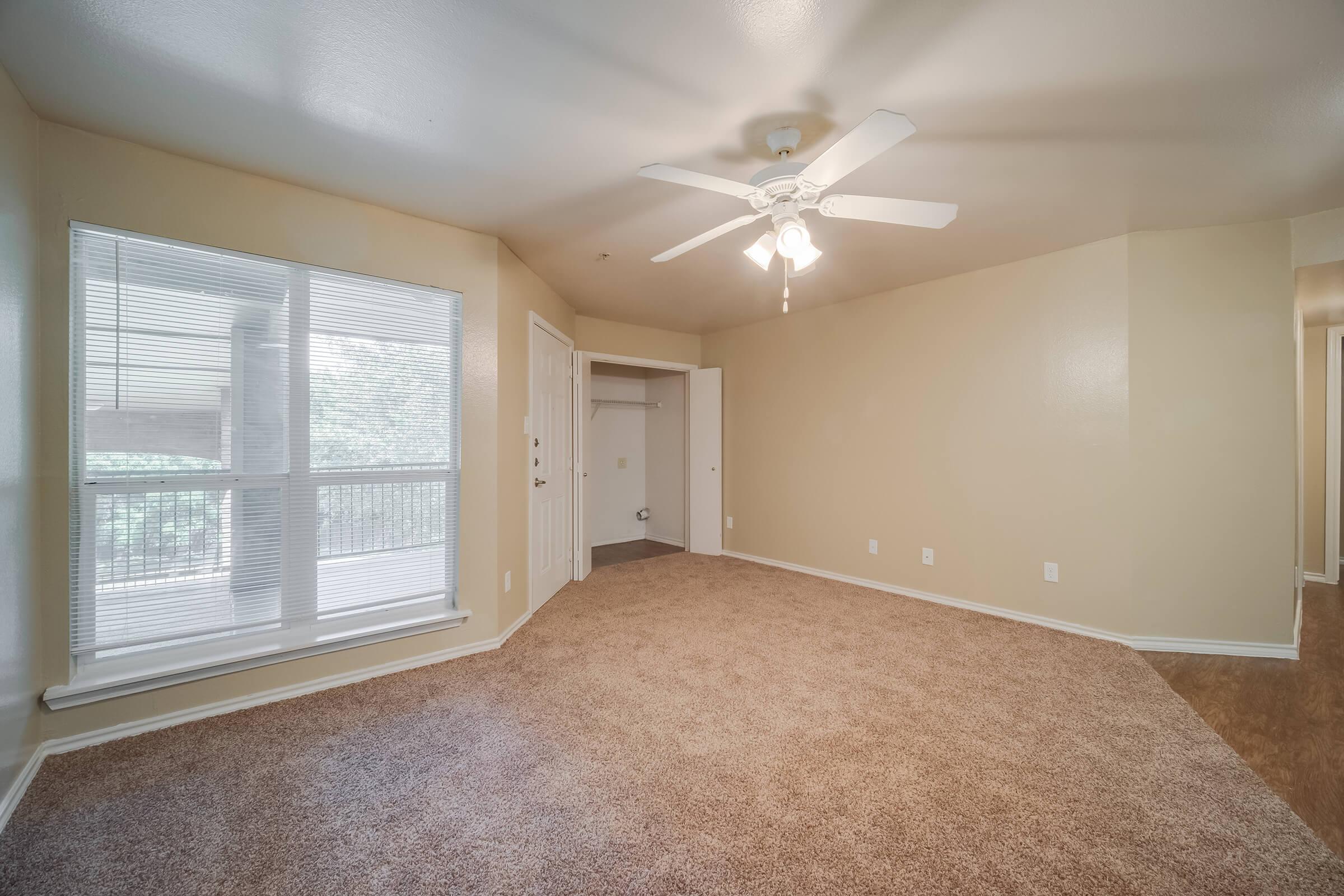 A spacious interior view of a room with light beige walls and a ceiling fan. The floor is covered with plush carpeting. A large window fills one corner with natural light, offering a view outside. An open closet is visible in the background, enhancing the sense of space.