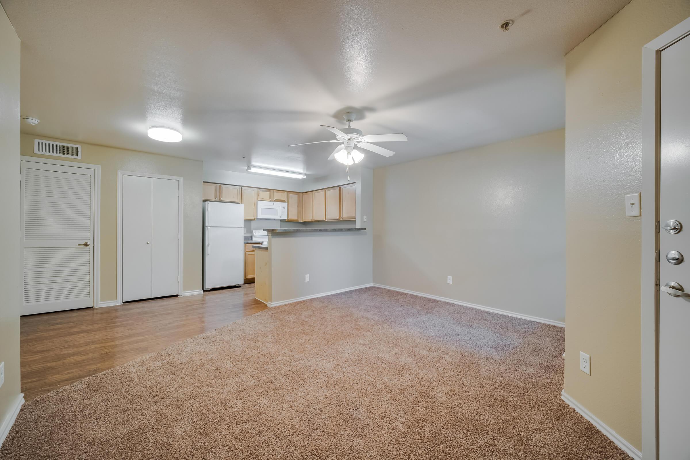 A spacious living area featuring light beige walls, a ceiling fan, and plush carpet. The open floor plan connects to a kitchen area with wooden cabinets and silver appliances. A door leads to the entrance, and the layout allows for natural light. The room has a welcoming atmosphere, ideal for cozy living.