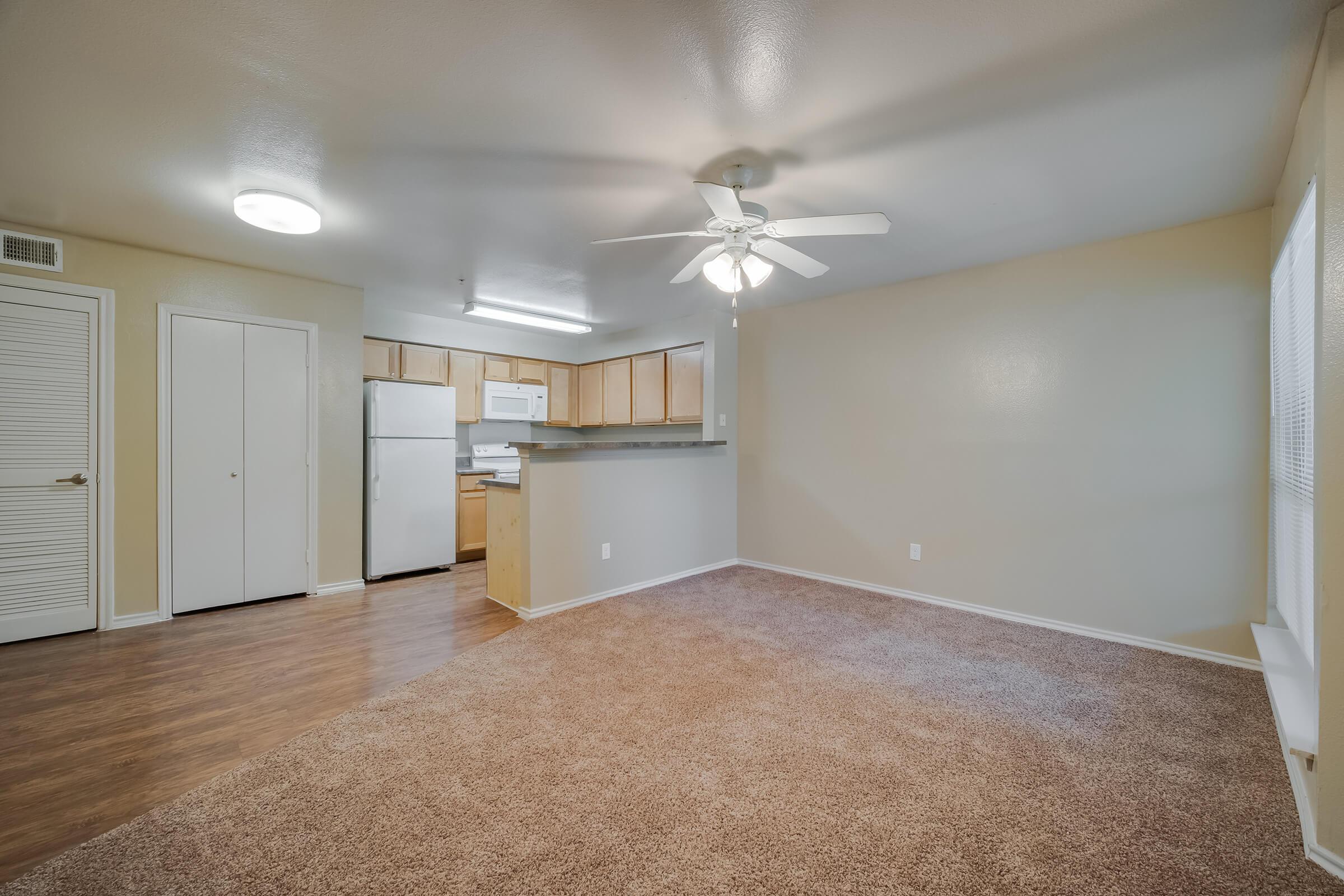 Interior view of a spacious living area featuring light brown carpet, a ceiling fan, and beige walls. A small kitchen area is visible in the background with wooden cabinets and a white refrigerator. Natural light enters through a window, enhancing the open and inviting atmosphere of the room.