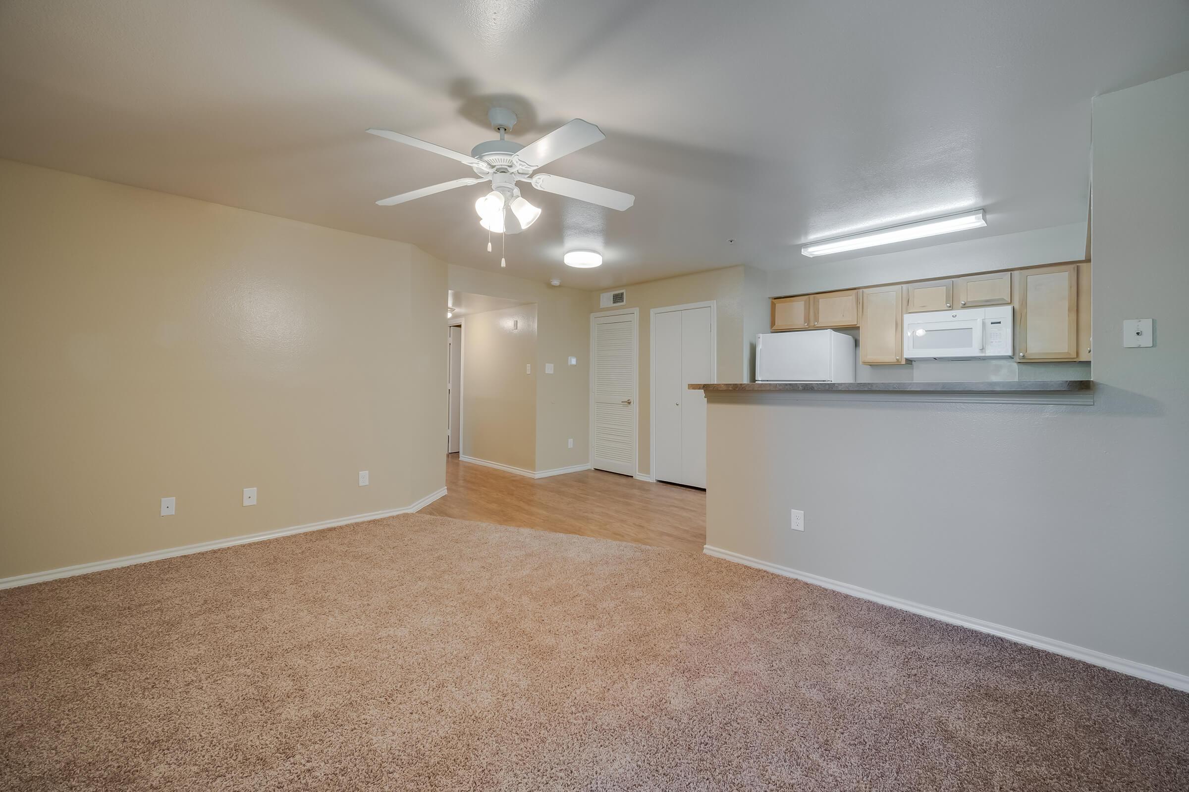 Spacious living area with light brown carpet, a ceiling fan, and neutral-colored walls. A kitchen with light wood cabinets is visible in the background, featuring a microwave and refrigerator. The layout leads to a hallway with closed doors, creating a cozy and functional space.