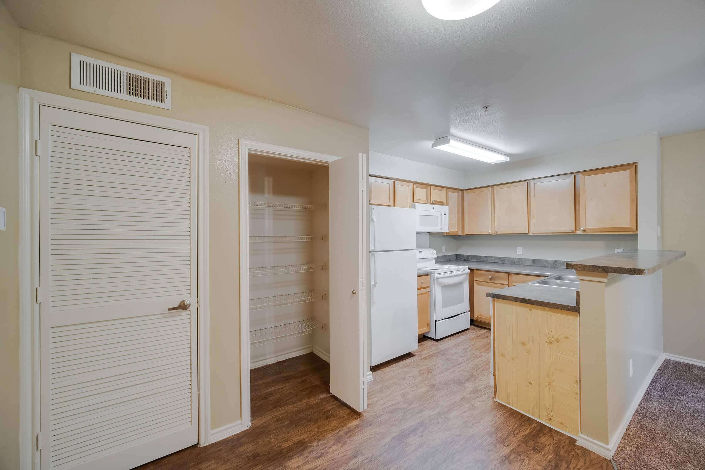 A roomy kitchen featuring wooden cabinets, a white refrigerator, and a stove. There’s an open pantry space with shelves on the left, and the kitchen is well-lit with overhead lighting. The floor is a wood-like laminate, creating a warm atmosphere.