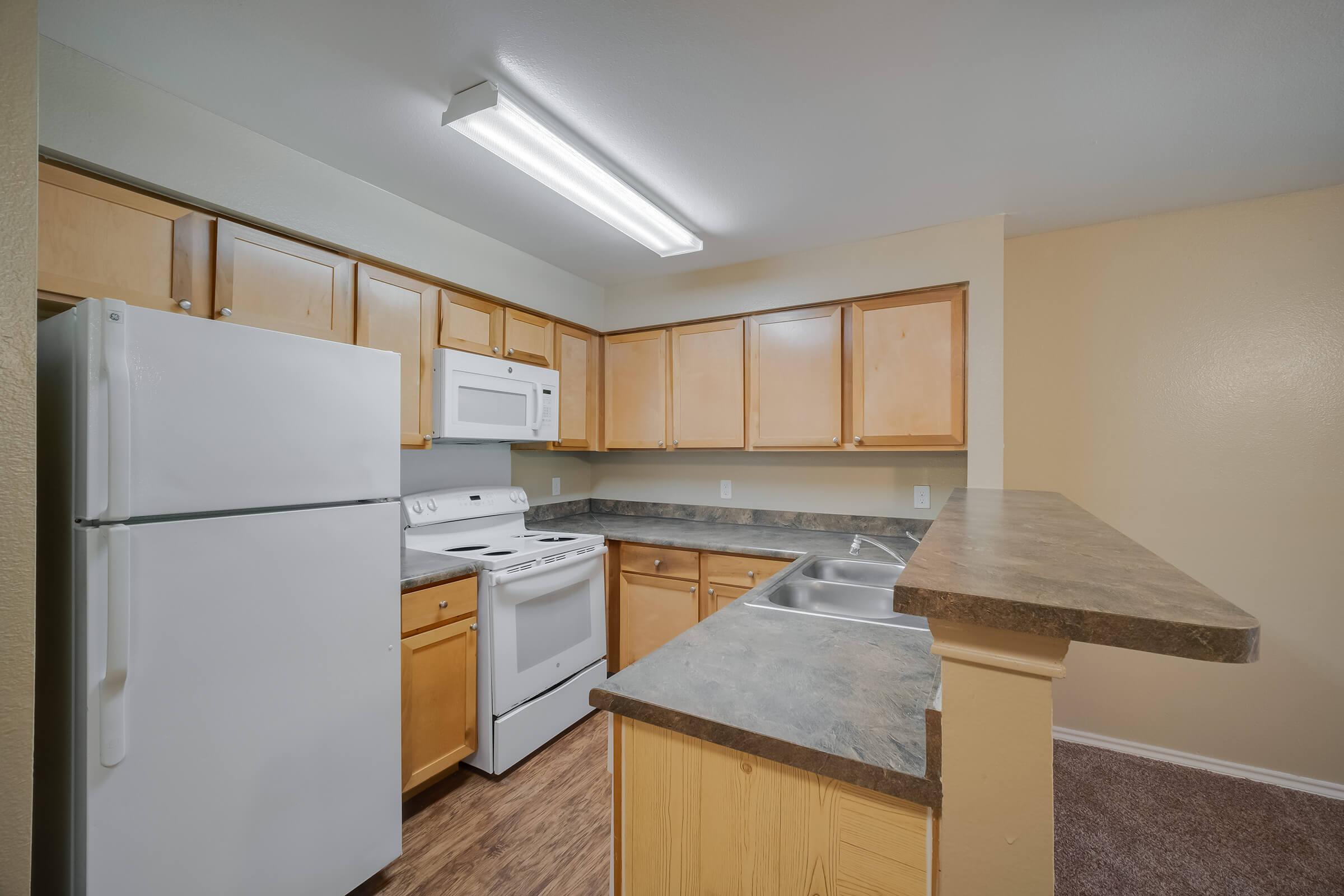 A modern kitchen featuring light-colored wooden cabinets, a white refrigerator, an oven, and a microwave. The countertops are dark with a sink, and the walls are painted a soft beige. Overhead, there is a bright overhead light fixture. The floor has a wood-like appearance, providing a cozy atmosphere.