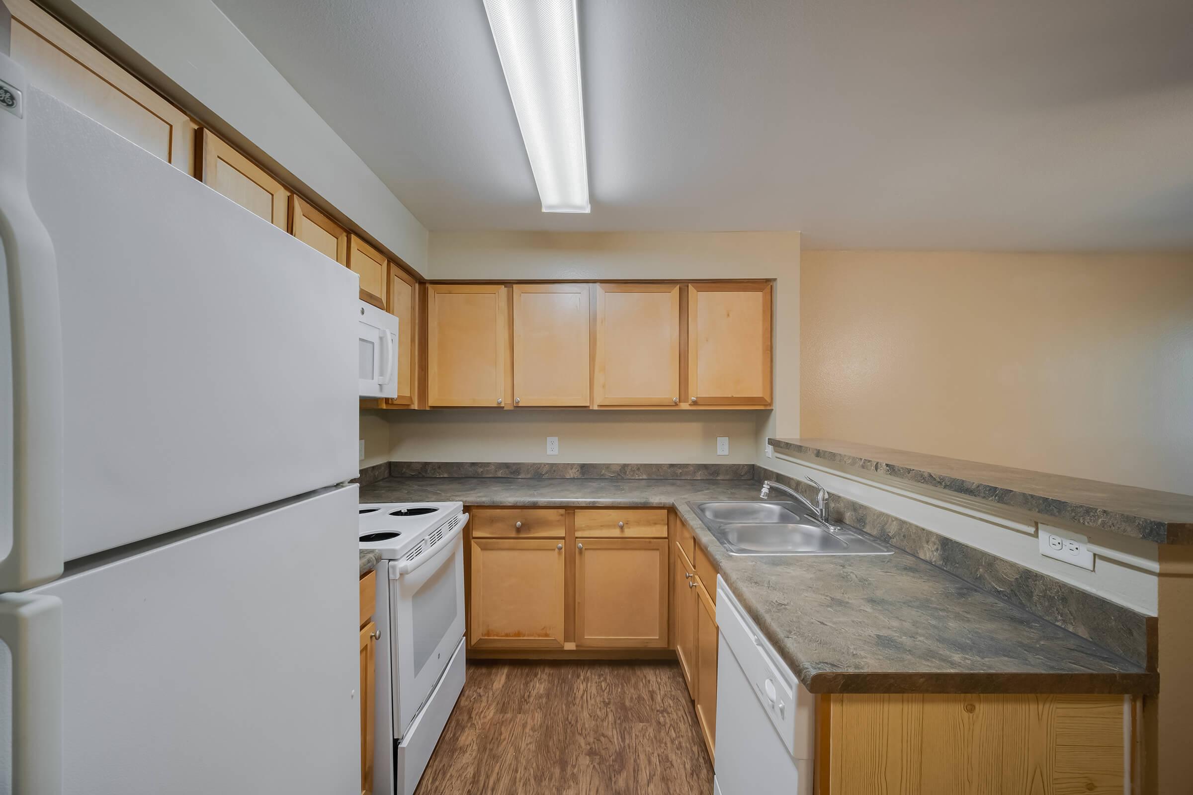 A modern kitchen featuring wooden cabinetry, a countertop, and essential appliances. Includes a white refrigerator, microwave, stove, oven, dishwasher, and dual sink. The walls are painted in a soft tone, and overhead fluorescent lighting illuminates the space, highlighting the clean and tidy layout.