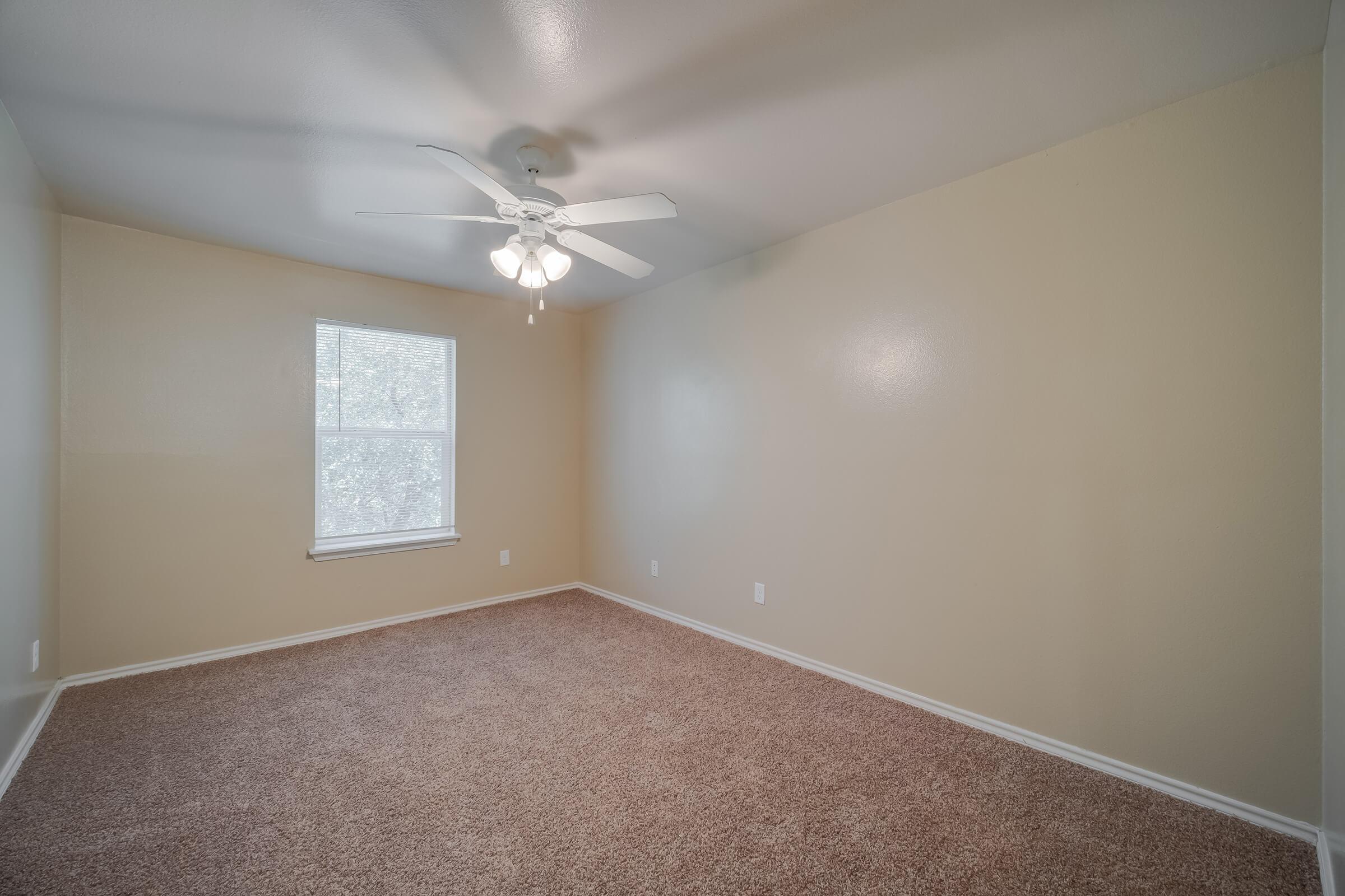 A bright, empty room featuring beige walls and a light brown carpet. It has a window with blinds letting in natural light and a ceiling fan with white blades. The space is uncluttered, making it suitable for various uses, with a clean and open atmosphere.