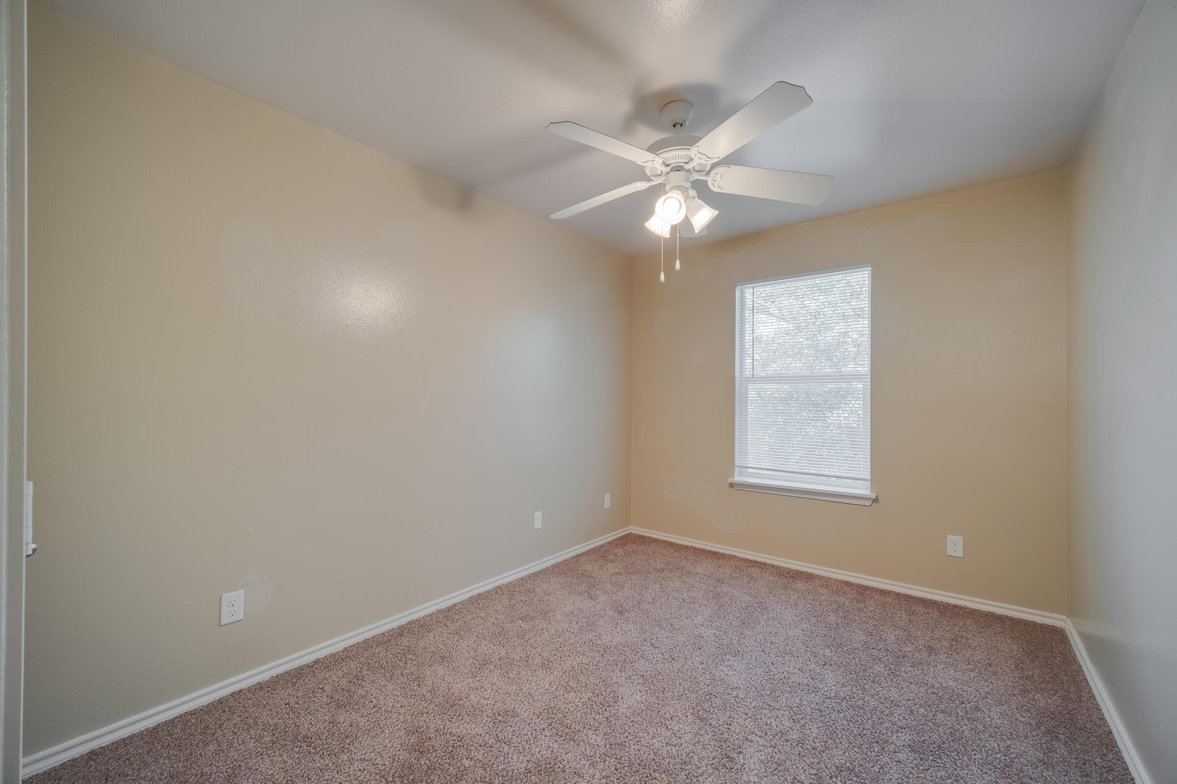 A vacant room with beige walls and carpeted flooring. It features a ceiling fan with three lights and a window that allows natural light to enter. The room is uncluttered, creating a spacious appearance, and has a simple, neutral aesthetic.