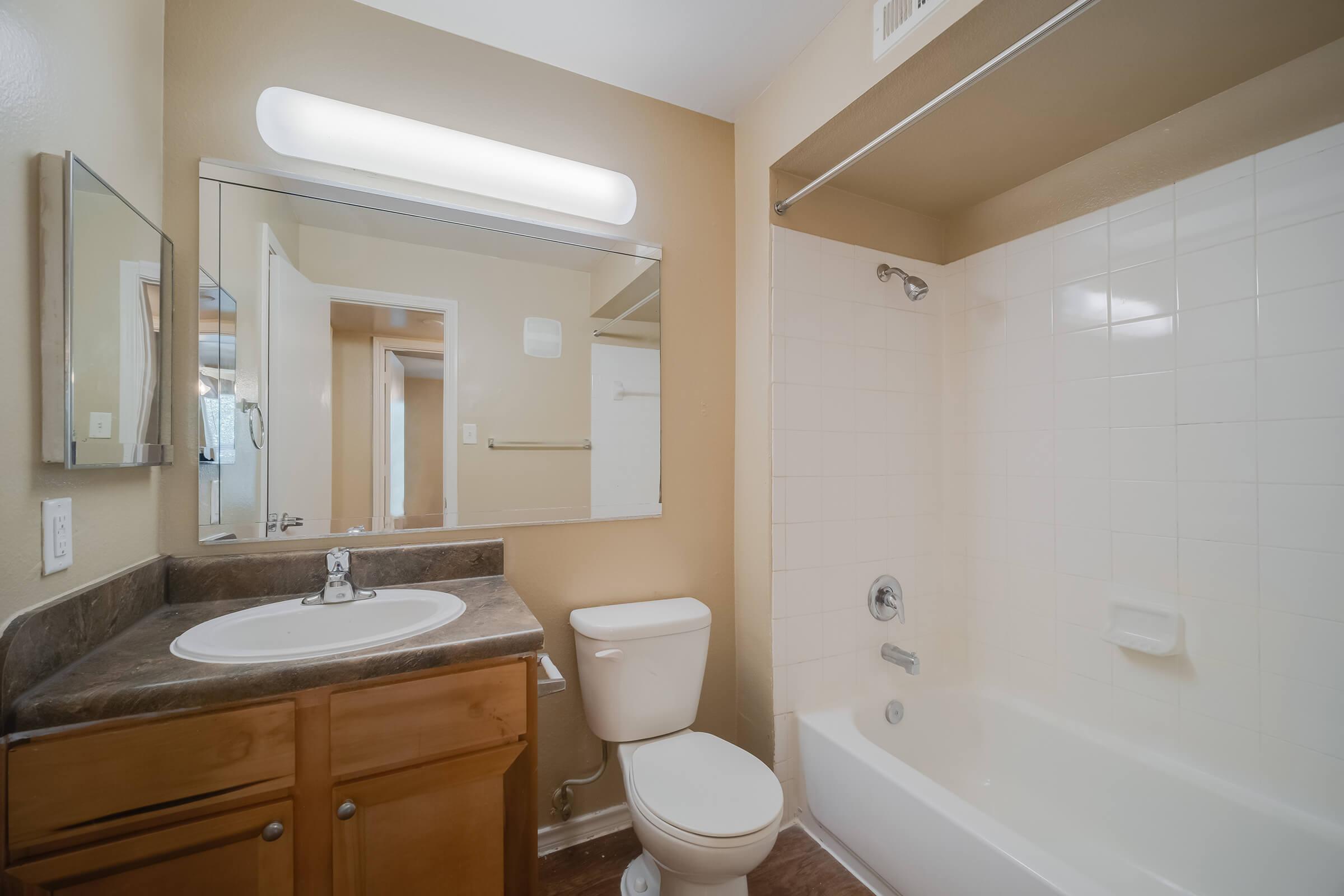 A clean, well-lit bathroom featuring a bathtub with a shower, a white toilet, and a sink with a wooden vanity. There is a large mirror above the sink and light fixtures on the wall. The walls are painted a light tan color, and the flooring appears to be laminate. A door leads to another room.