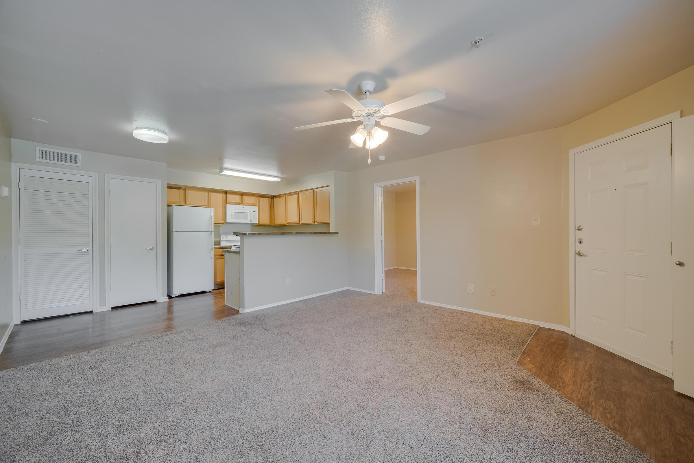 A spacious living area featuring beige walls and plush carpet, with a ceiling fan overhead. An open kitchen is visible in the background, showcasing wooden cabinetry and modern appliances. The entrance door is on the right, leading to a hallway and additional rooms. Natural light fills the space.