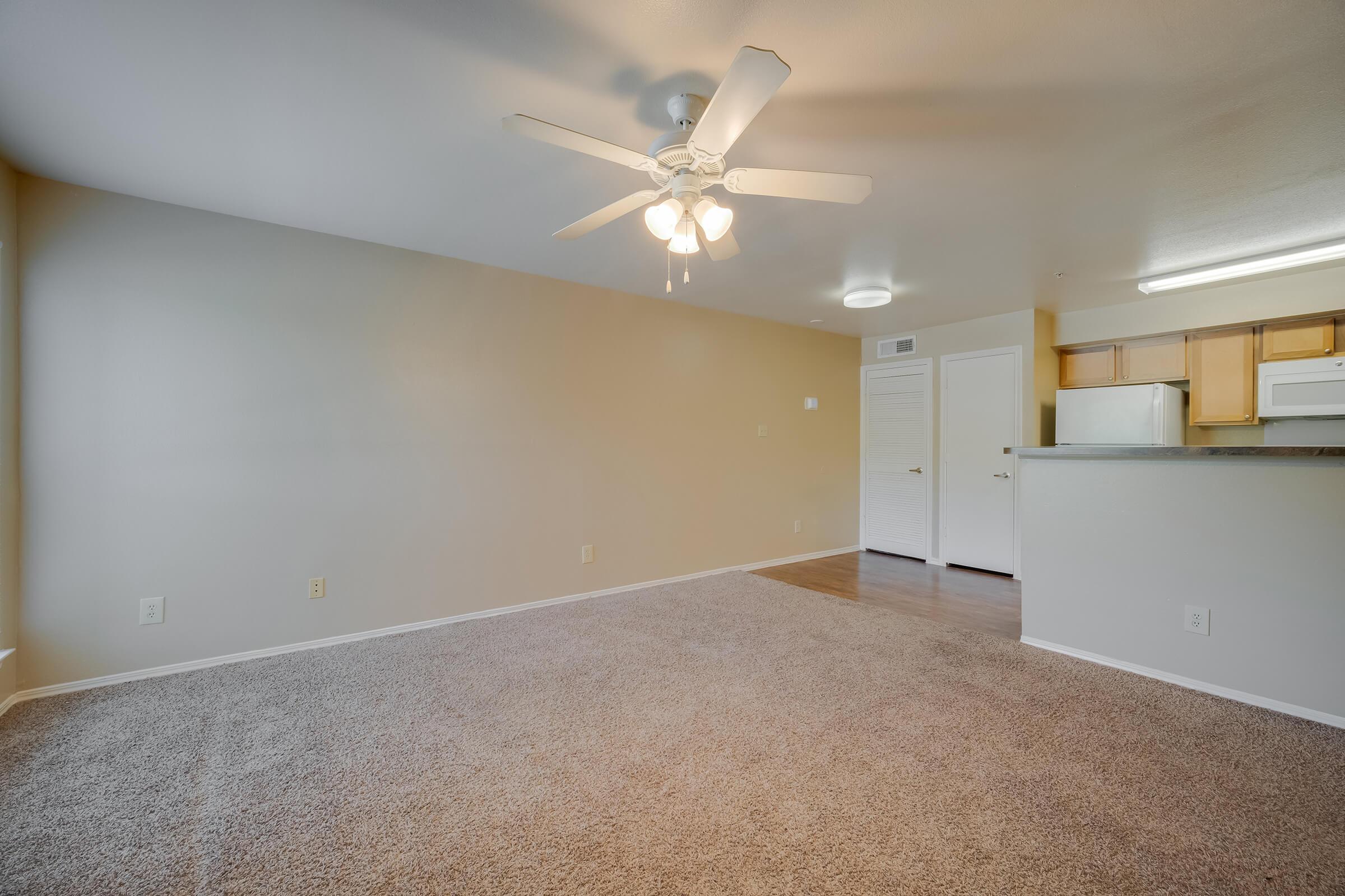 Interior view of a spacious living room featuring light beige walls, a ceiling fan with four lights, and plush carpet. The open layout connects to a kitchen area with a bar counter and white appliances. Natural light enters through a window, enhancing the warm and inviting atmosphere.