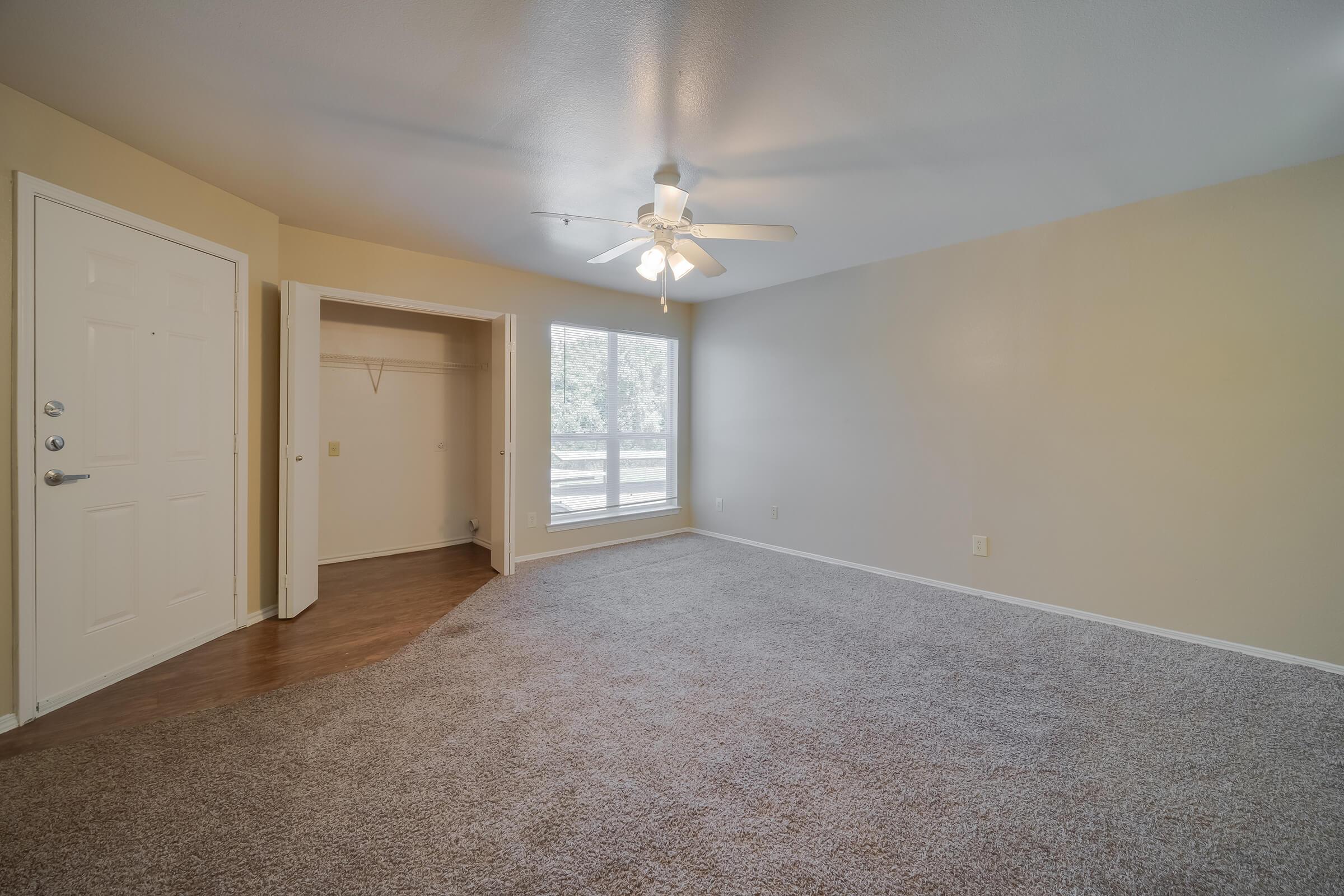 Spacious room with a light-colored wall and plush carpet, featuring a ceiling fan and a large window for natural light. A closet is visible near the entrance, and the door leads outside. The overall atmosphere is bright and inviting, suitable for various uses like a living area or bedroom.