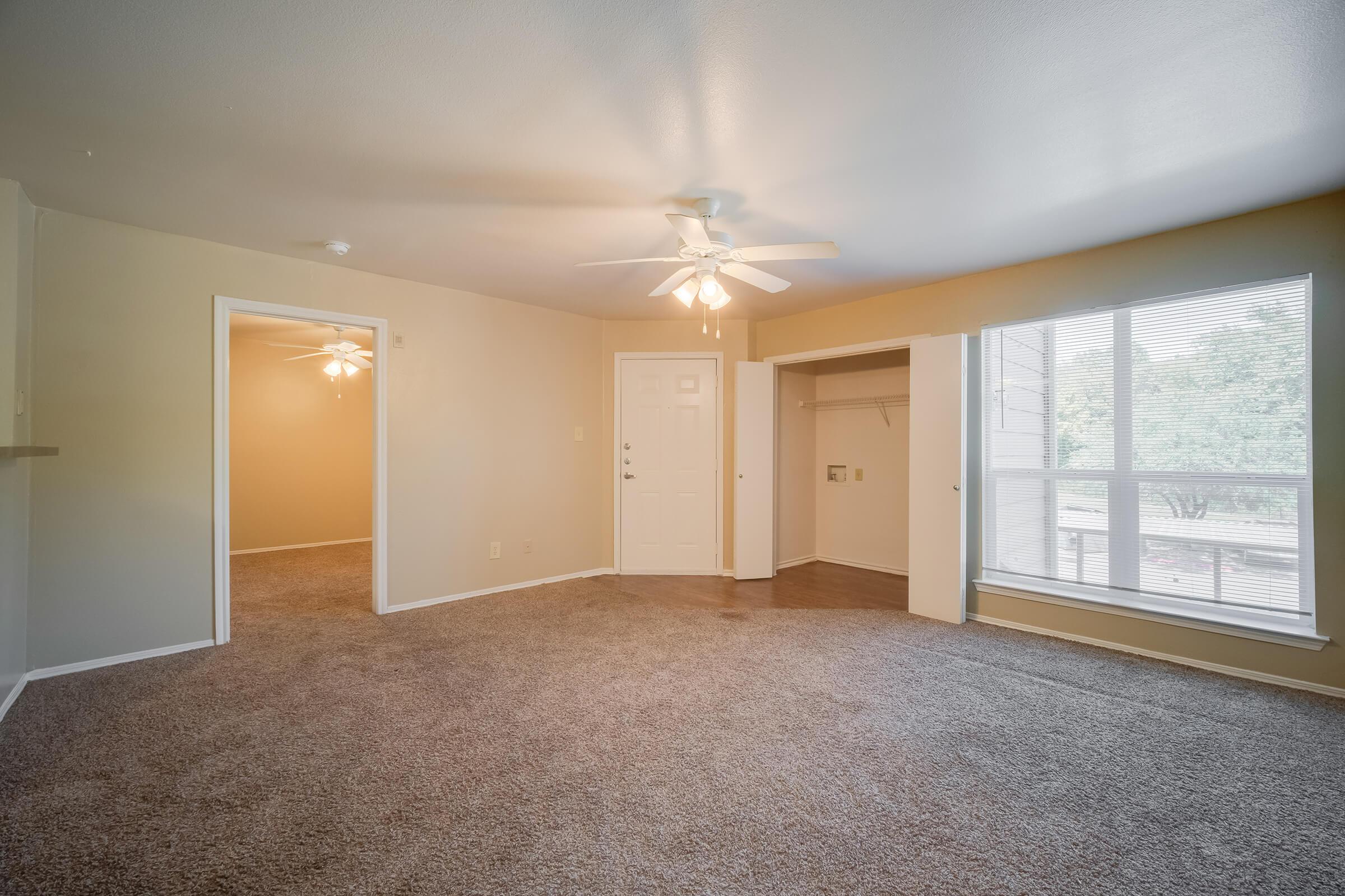 Spacious living room with beige walls and carpet, featuring a ceiling fan and large window. The room leads to a hallway with an open door, revealing a closet. Natural light pours in, creating a warm ambiance, while a front door is visible, enhancing the inviting feel of the space.