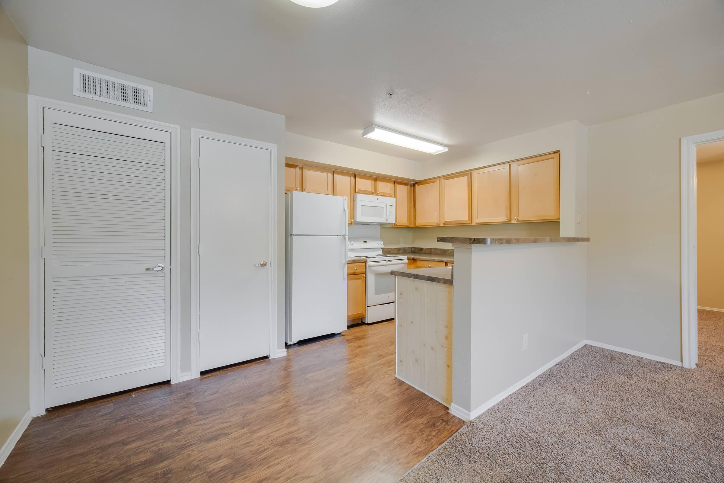 Bright and spacious kitchen area featuring wooden cabinets, a white refrigerator, a stove, and a countertop. The room has an open layout with light-colored walls and hardwood flooring. A doorway leads to a carpeted area, indicating a living space or dining room adjacent to the kitchen.