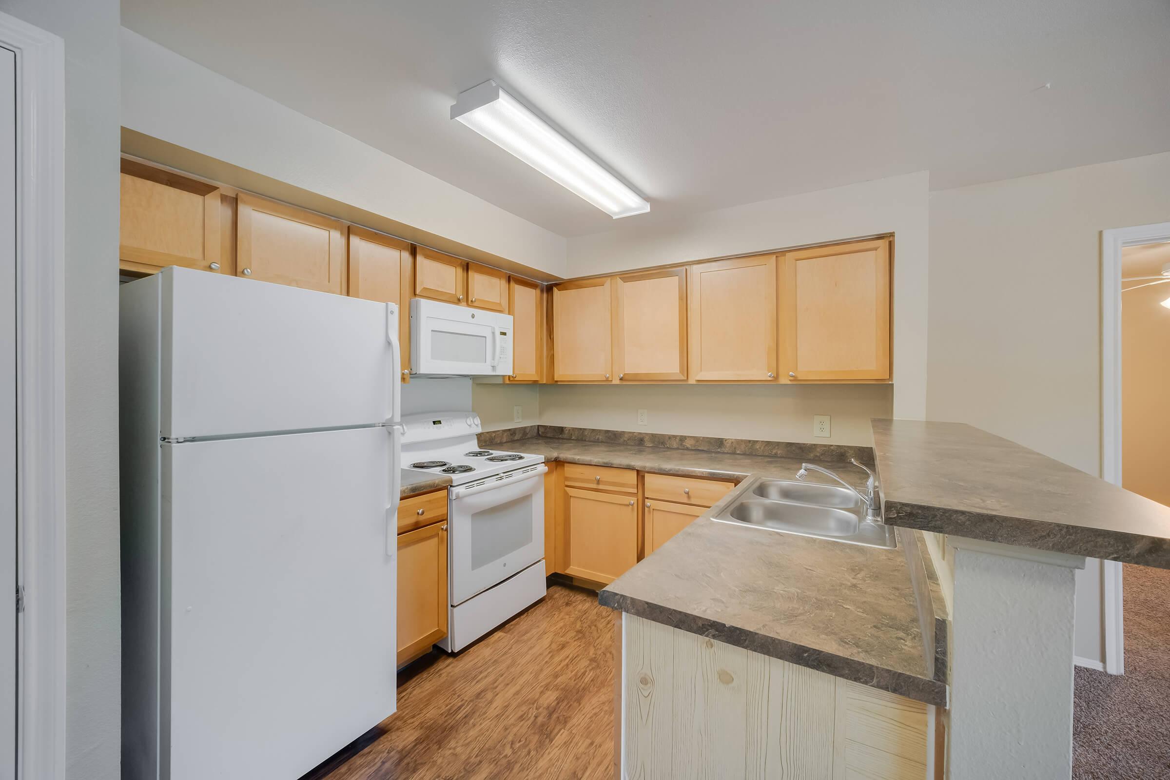 A modern kitchen featuring light wood cabinetry, a white refrigerator, a white microwave, and a white stove. Countertops in a dark tone complement the cabinetry. There are double sinks in the bar area, and the space has bright, overhead lighting. A neutral-colored wall completes the inviting atmosphere.