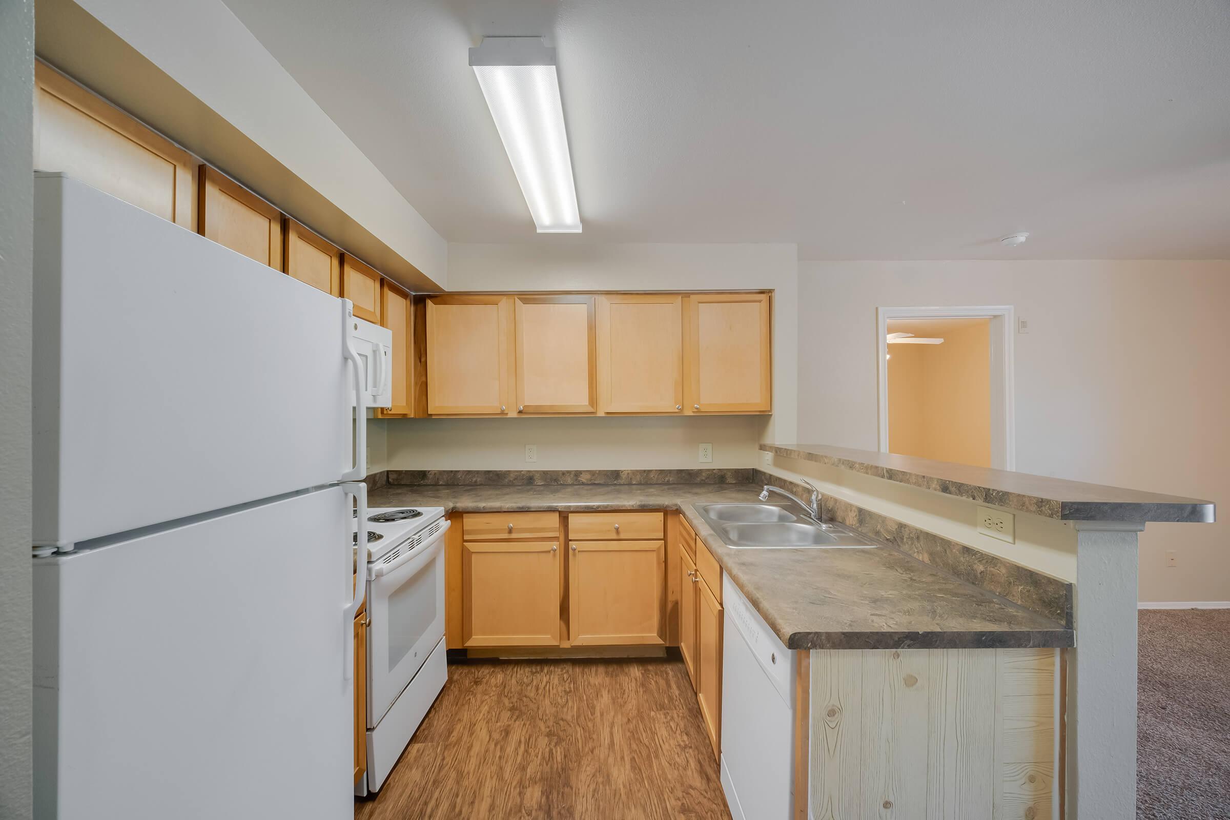 A spacious kitchen featuring light wood cabinets, a white refrigerator, a stove, and a sink. The countertops are dark and the flooring is a wood-like laminate. A bright overhead light enhances the openness of the space. The kitchen flows into a neighboring area with neutral-colored walls.