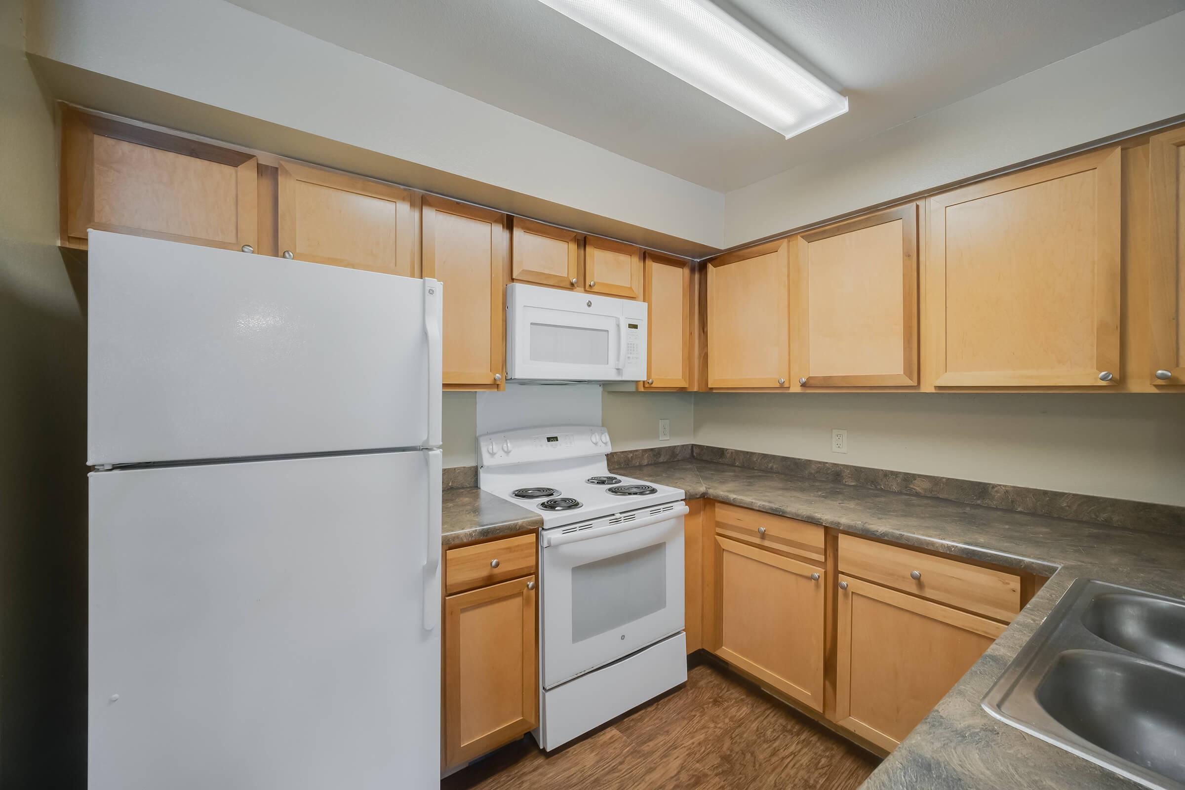 Modern kitchen featuring light wood cabinets, a white refrigerator, a white stove with an oven, and a built-in microwave. The countertop is dark, complementing the cabinetry. The space is well-lit with overhead fluorescent lighting.