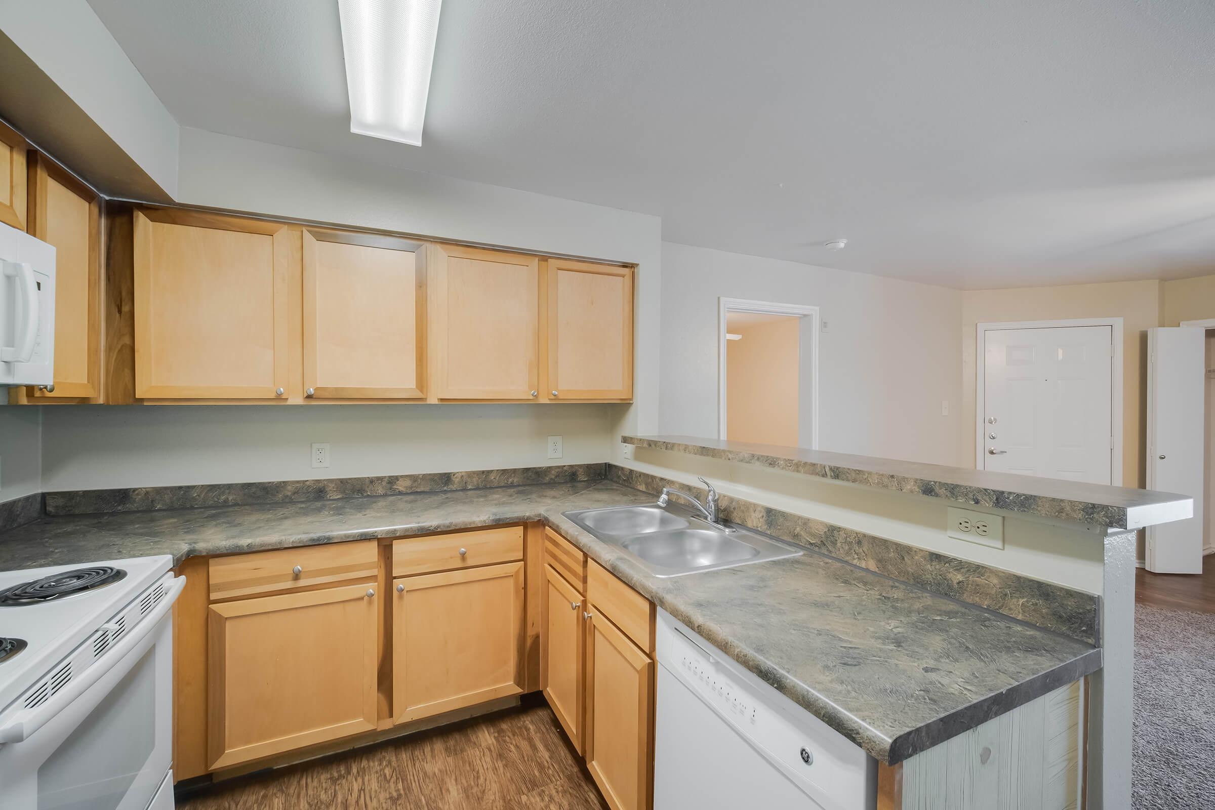 A well-lit kitchen with wooden cabinets, a double sink, and modern appliances including a stove and dishwasher. The countertops are a dark color, and the space opens into a light-colored living area. The flooring appears to be laminate, contributing to a clean and functional design.