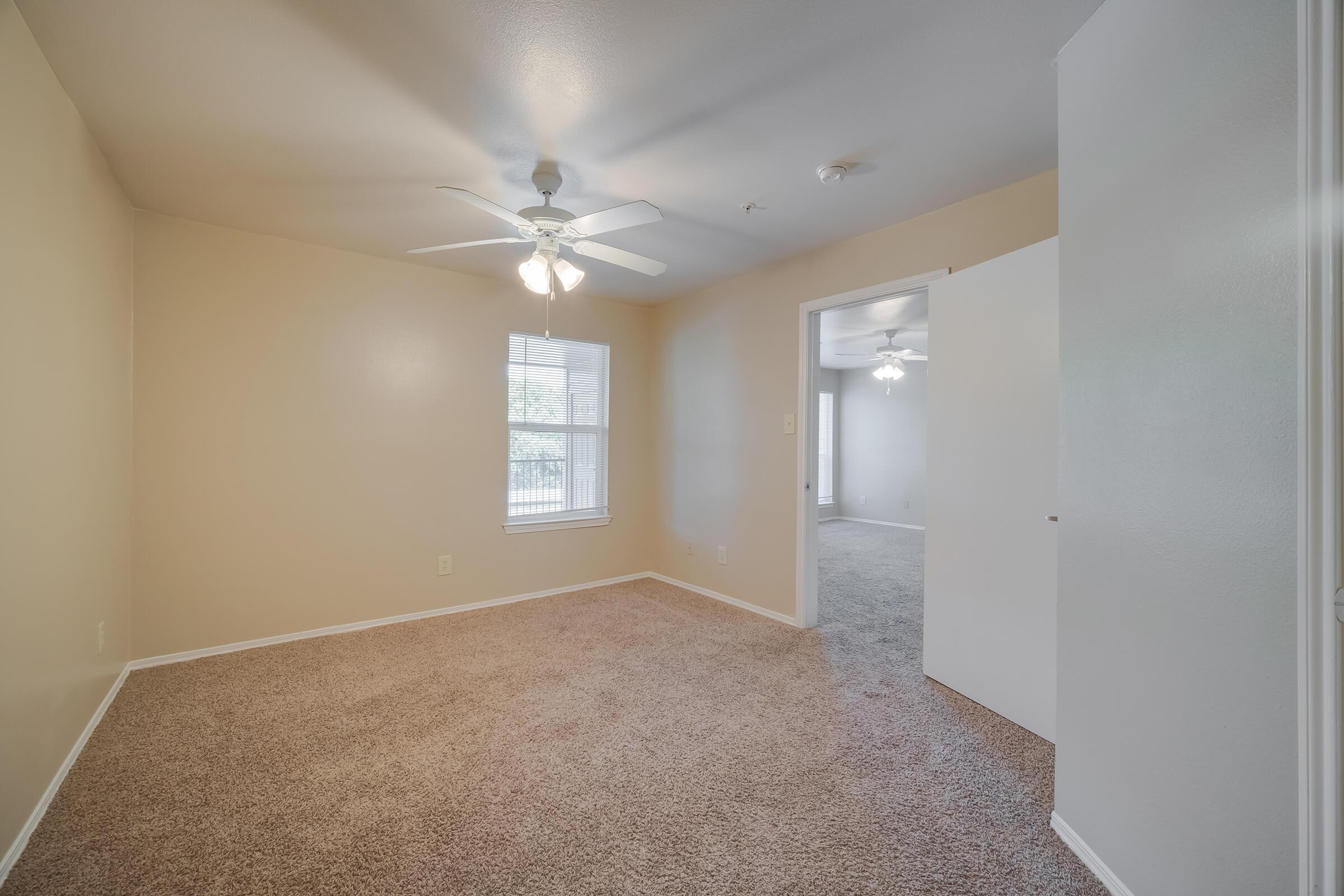 A well-lit, empty room featuring beige walls and a light brown carpet. It has a ceiling fan and a window letting in natural light. An open doorway leads to another room, with a similar color scheme, visible in the background. The overall atmosphere is clean and spacious, ideal for personal use or decoration.