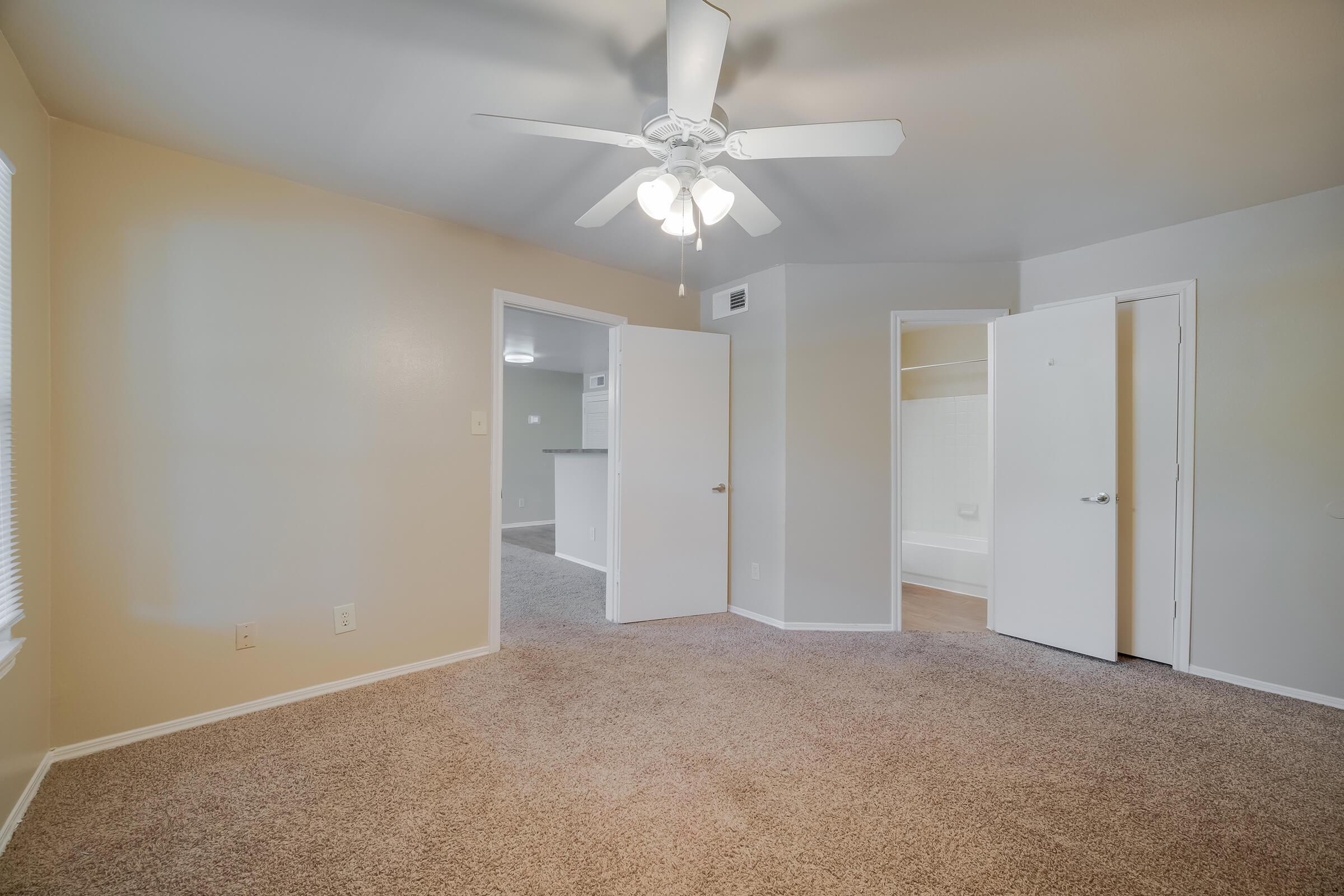 A well-lit room featuring neutral-colored walls and plush carpet. It has a ceiling fan with white blades, two doors leading to other areas, and a window allowing natural light. One door is slightly open, revealing a bathroom area. Overall, the space appears clean and inviting.