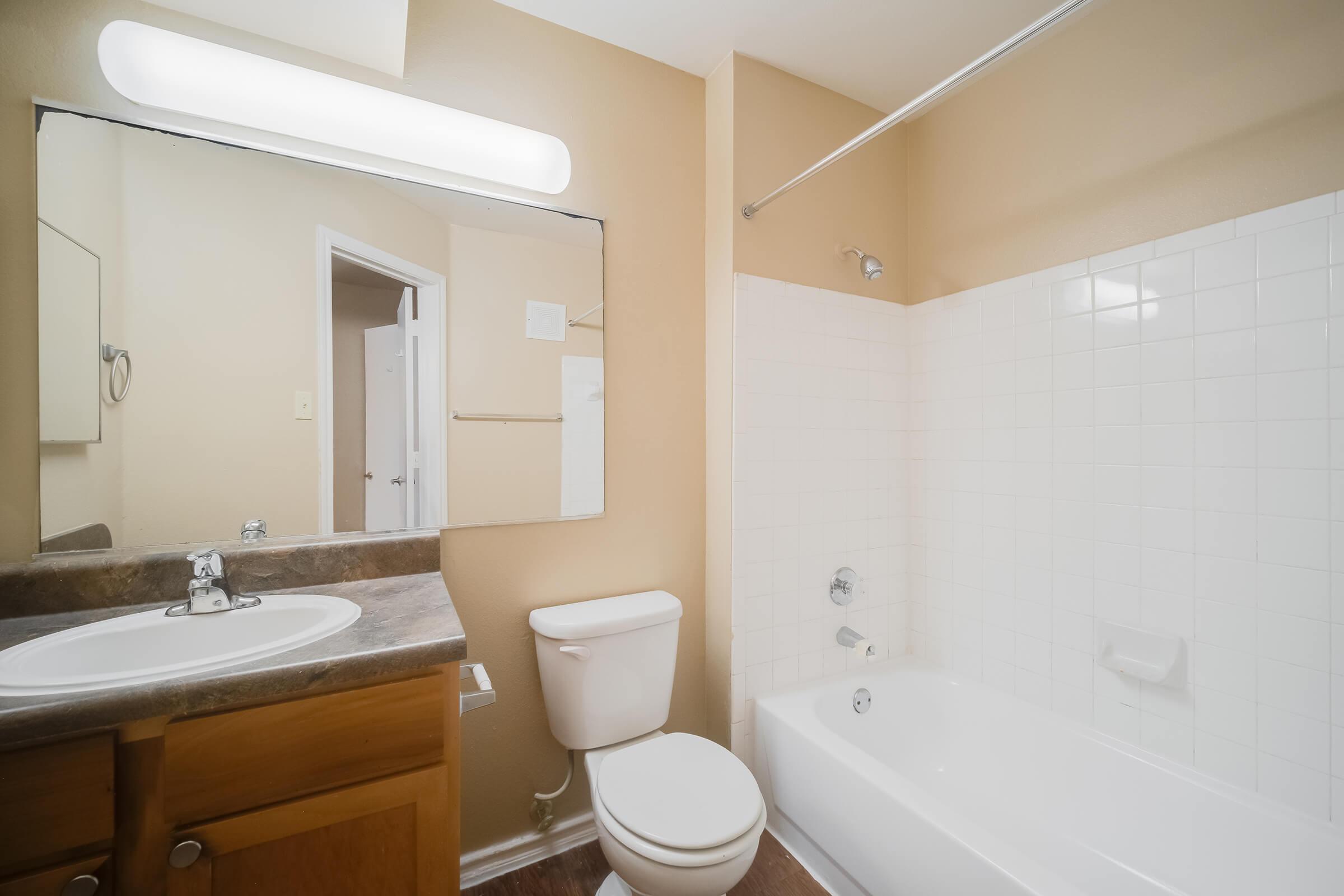 A clean and simple bathroom featuring a white bathtub, a toilet, and a sink with a wooden cabinet. The walls are painted in a soft beige color, and there is a large mirror above the sink. A fluorescent light is mounted above the mirror, illuminating the space. The bathroom appears well-maintained and functional.