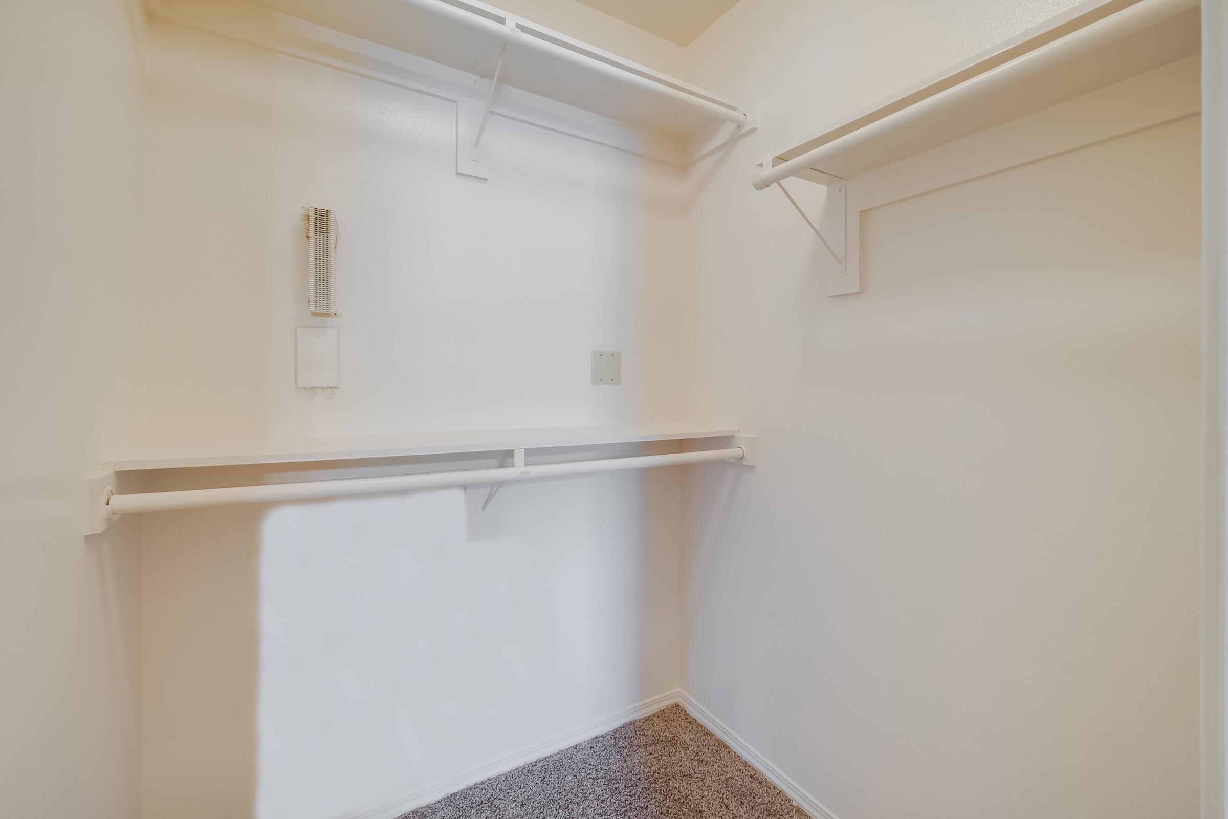 Empty closet with light-colored walls, two shelves mounted on the sides, and a soft carpeted floor. The space is uncluttered, providing ample room for storage or organization. There is a small control panel or device on the wall, but the overall appearance is minimalistic and tidy.