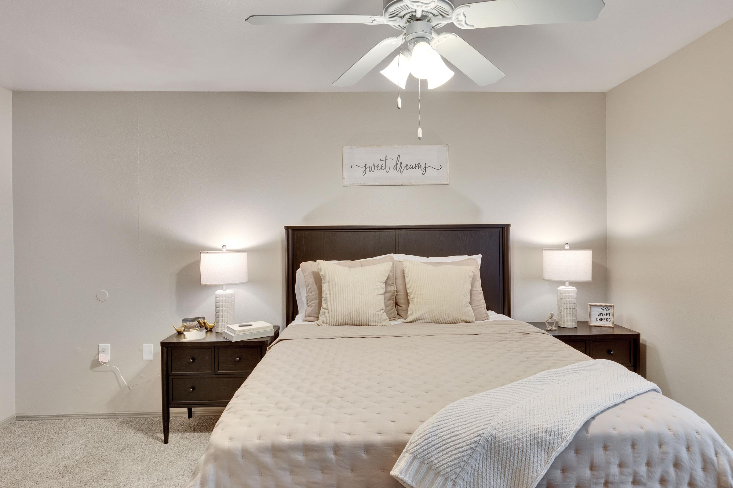 A cozy bedroom featuring a queen-sized bed with beige bedding, two nightstands with lamps on either side, a ceiling fan, and a decorative wall sign. The walls are painted in a soft hue, creating a calm atmosphere. A light throw blanket is draped over the bed for added comfort.