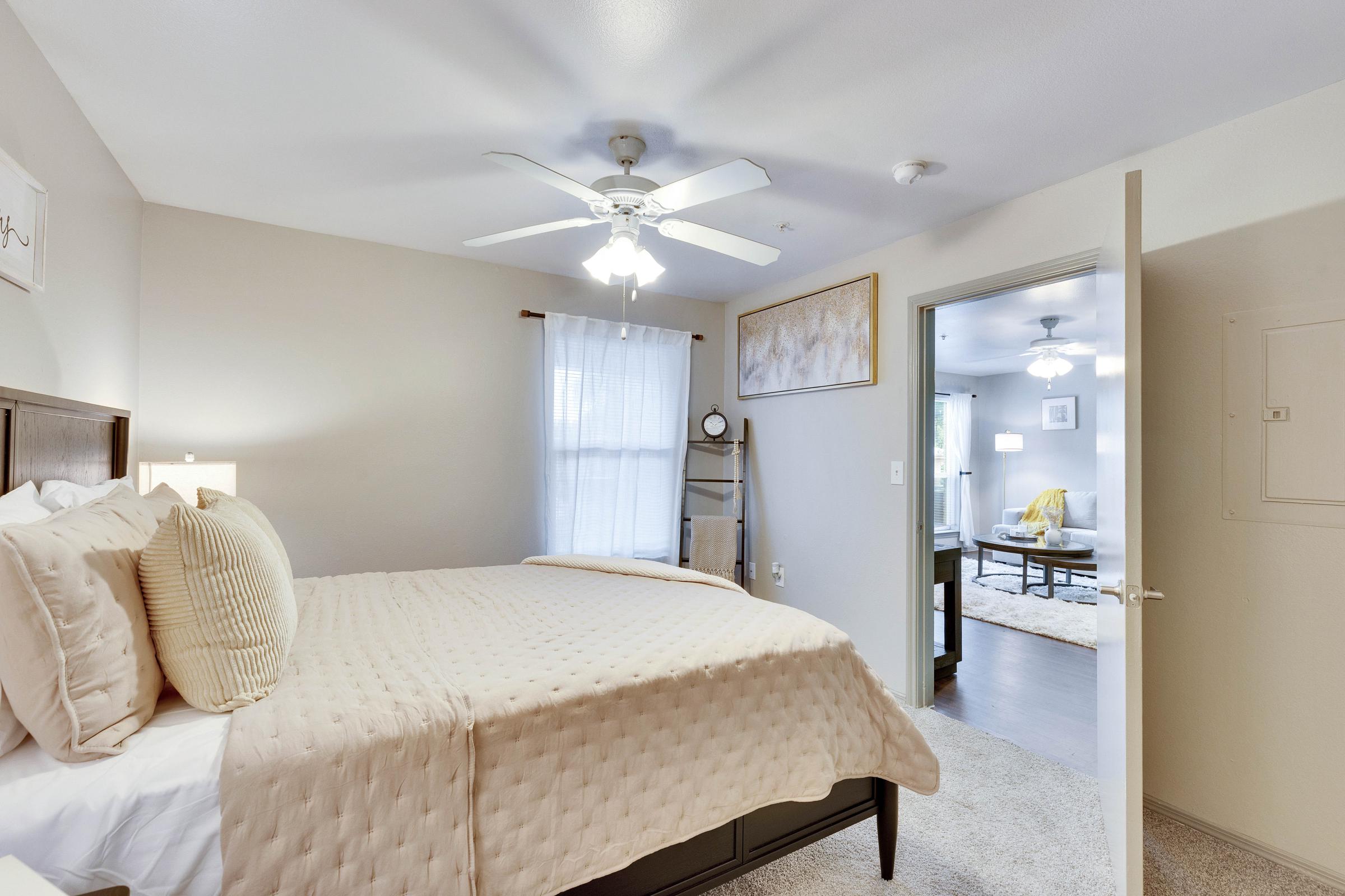 Cozy bedroom featuring a large bed with beige bedding, two bedside lamps, and a ceiling fan. A doorway leads to another room, while a window lets in natural light. The decor is neutral and inviting, creating a warm atmosphere in the space.