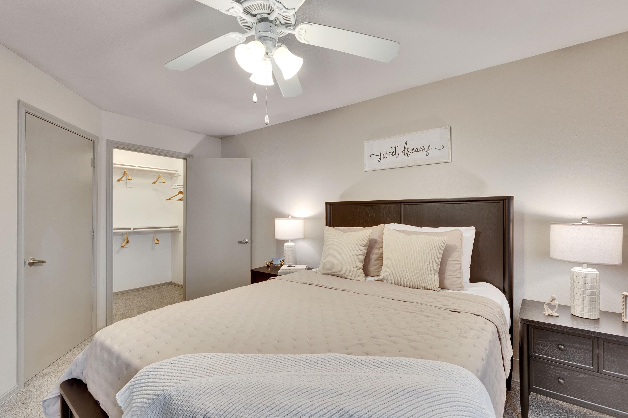 A cozy bedroom featuring a queen-sized bed with light brown bedding, two bedside tables with lamps, and a wall-mounted sign that reads "sweet dreams." A ceiling fan hangs above the bed, and a closet is visible in the background with wooden hangers. The wall and carpet are in neutral tones, creating a warm atmosphere.