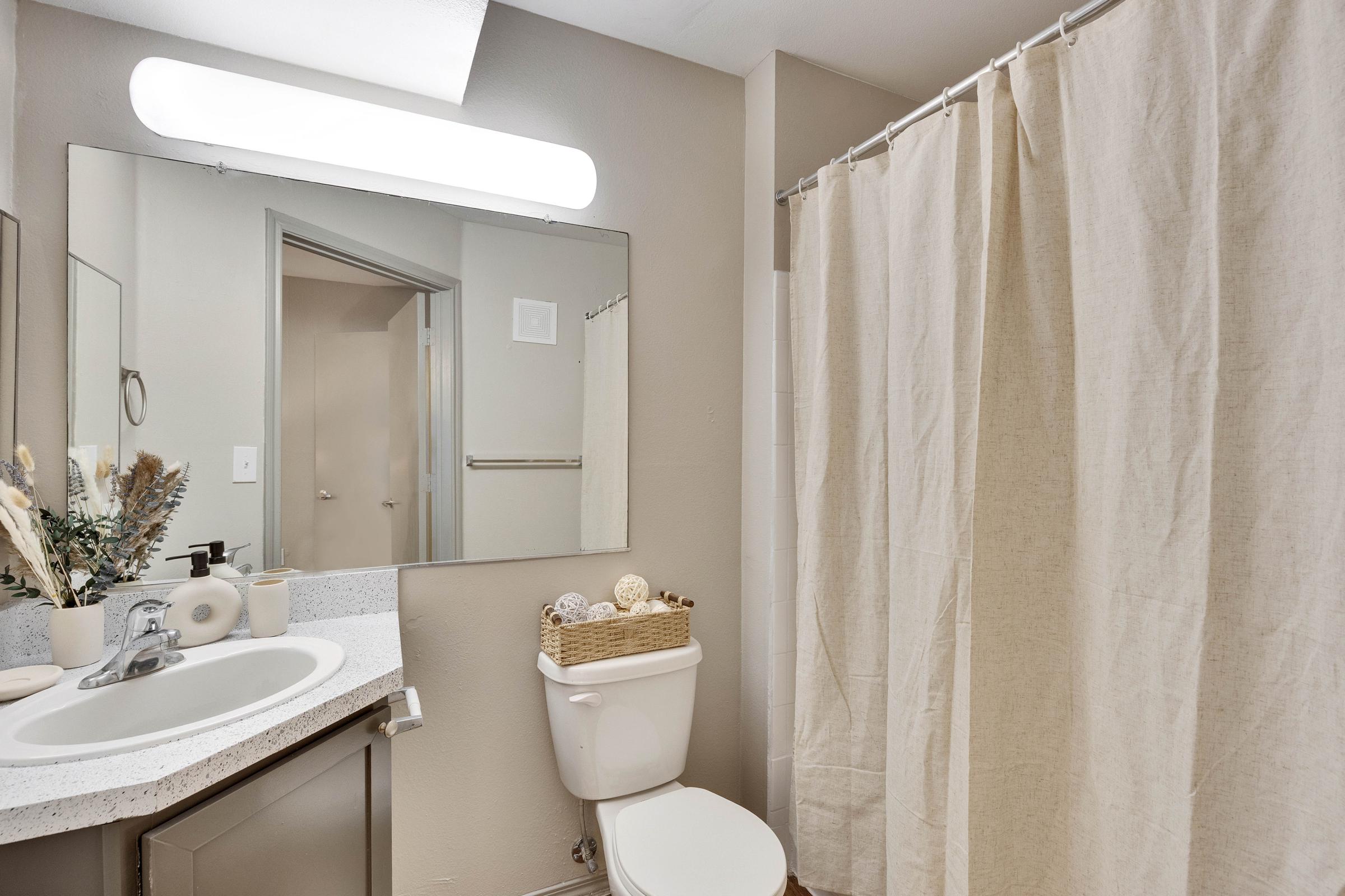 A clean and modern bathroom featuring a white shower curtain, a countertop with sink, and decorative items. A large mirror reflects the space, and there's a towel rack visible in the background. The color scheme is neutral, creating a calm atmosphere.
