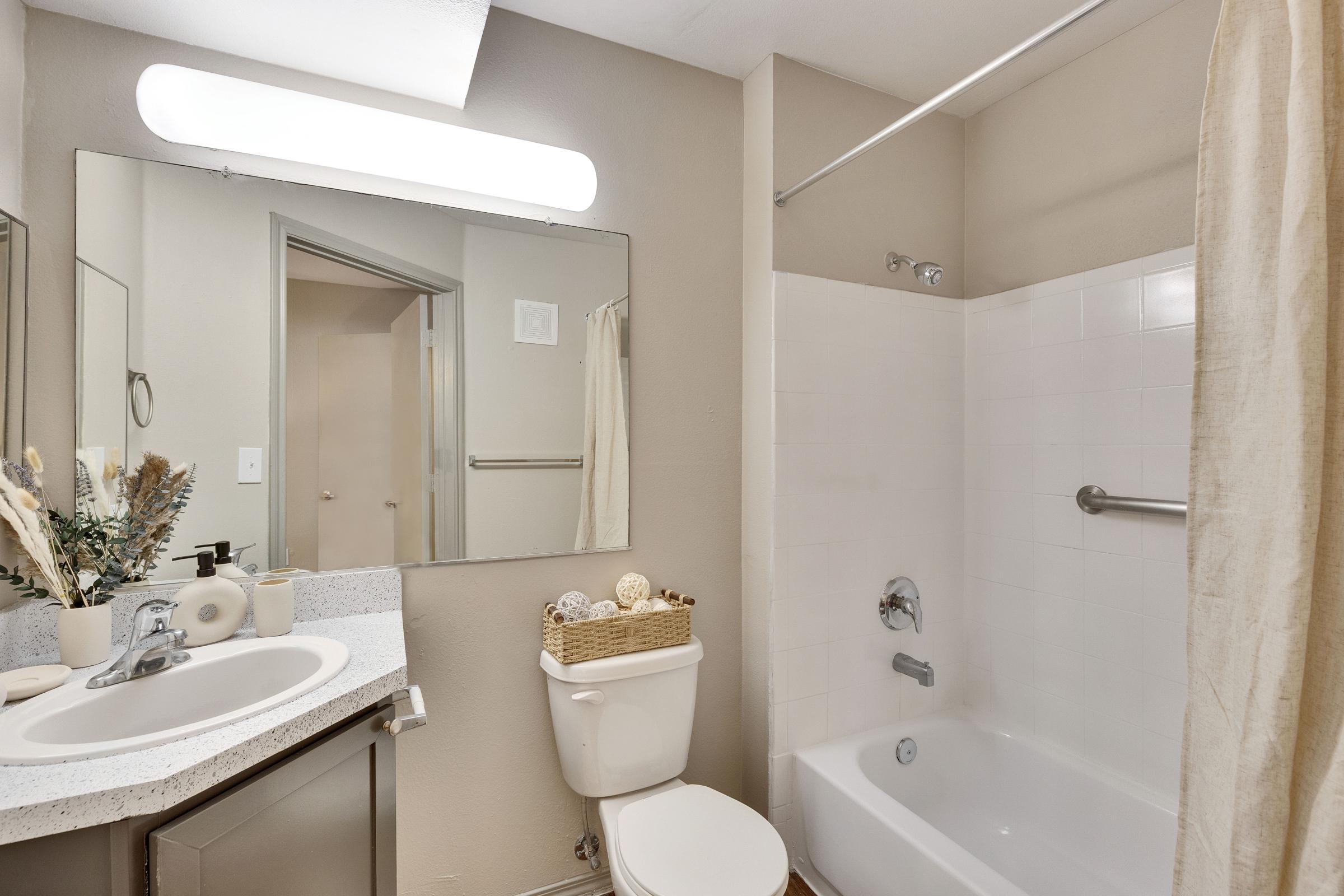 A clean and modern bathroom featuring a bathtub with a shower, a white toilet, and a sleek vanity with a countertop. The walls are painted light beige, and there’s a mirror above the sink. A small basket is placed on the vanity, adding a touch of decor. Natural light illuminates the space.