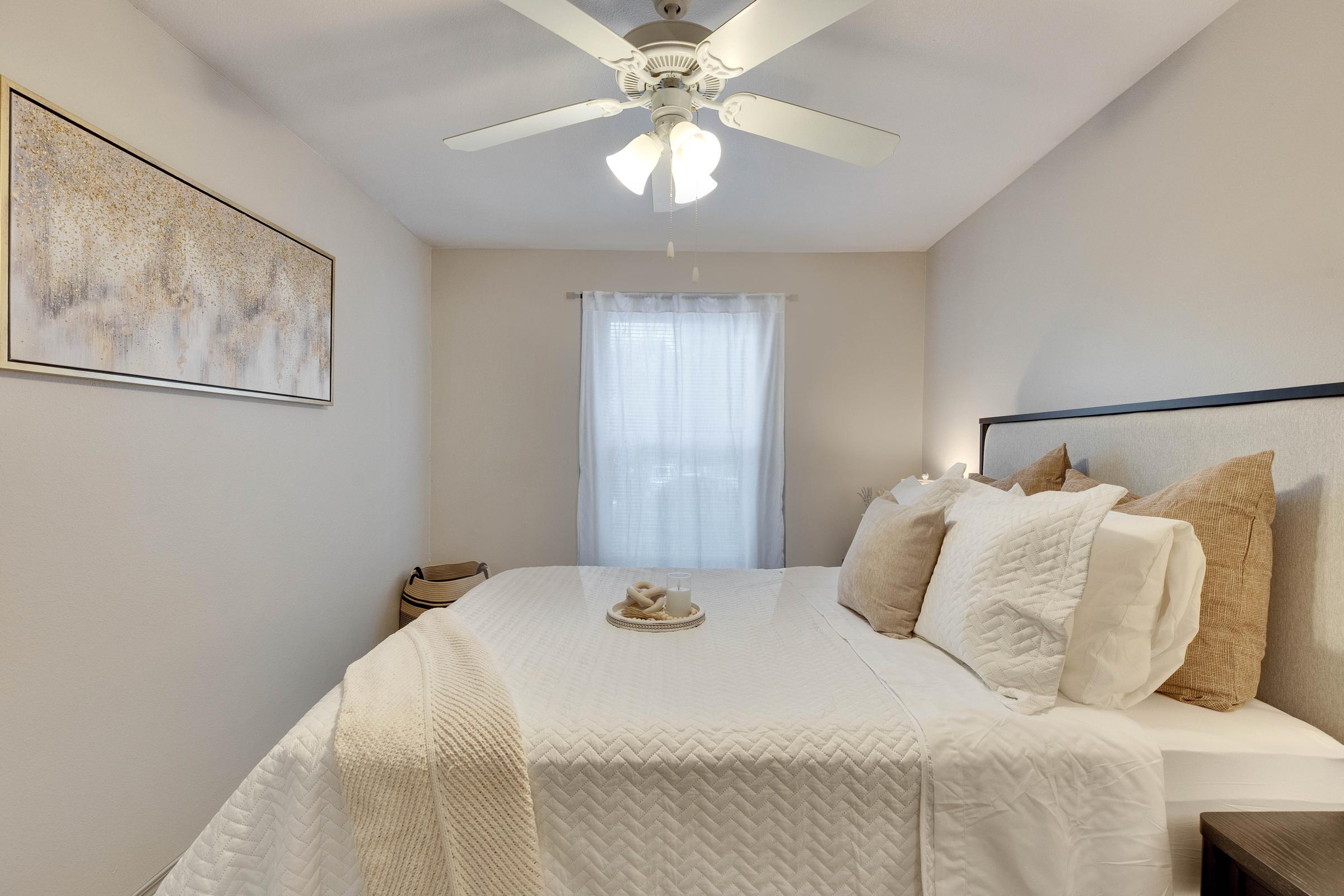 A cozy bedroom featuring a neatly made bed with beige and white bedding, adorned with decorative pillows. The room has light-colored walls, a ceiling fan, and a window covered with sheer curtains, allowing natural light to filter in. A piece of wall art and a soft throw are also present, adding warmth to the space.