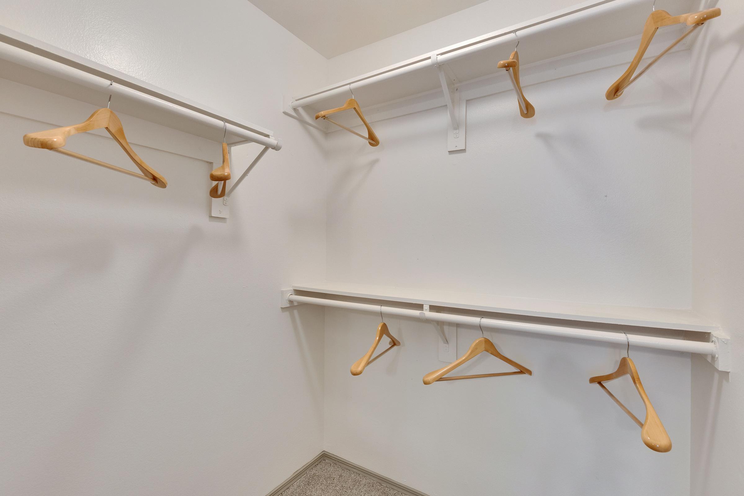 Empty closet with white walls and two rows of white hanging rods. Several wooden hangers are suspended from the rods, with some also placed on a shelf. The floor is covered in grey carpeting.