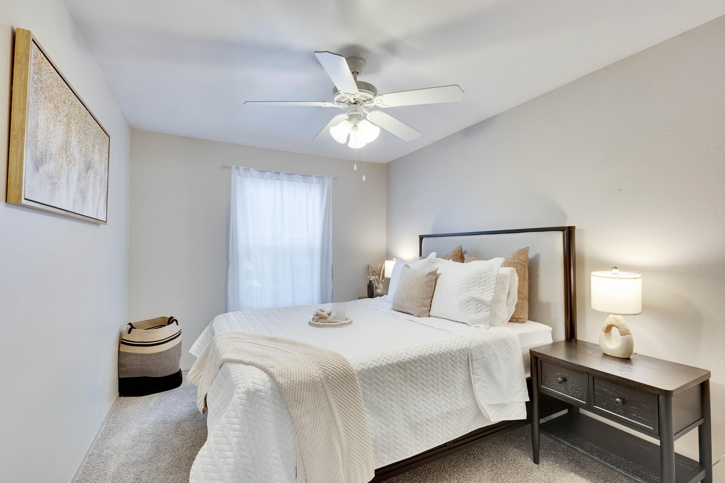 A cozy bedroom featuring a queen-sized bed with white bedding, decorative pillows, and a light throw. There's a ceiling fan, a window with sheer curtains, a bedside lamp on a dark wooden nightstand, and a woven basket beside the bed, enhancing the warm and inviting atmosphere.