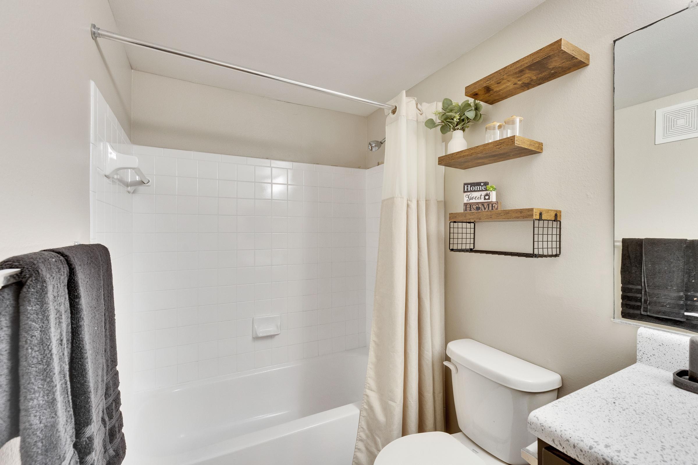 A clean bathroom featuring a white bathtub with a shower curtain, a wooden shelf with decorative items, and a mirror above a sink. There are towels hanging on a rack and a toilet nearby, creating a modern and organized space.