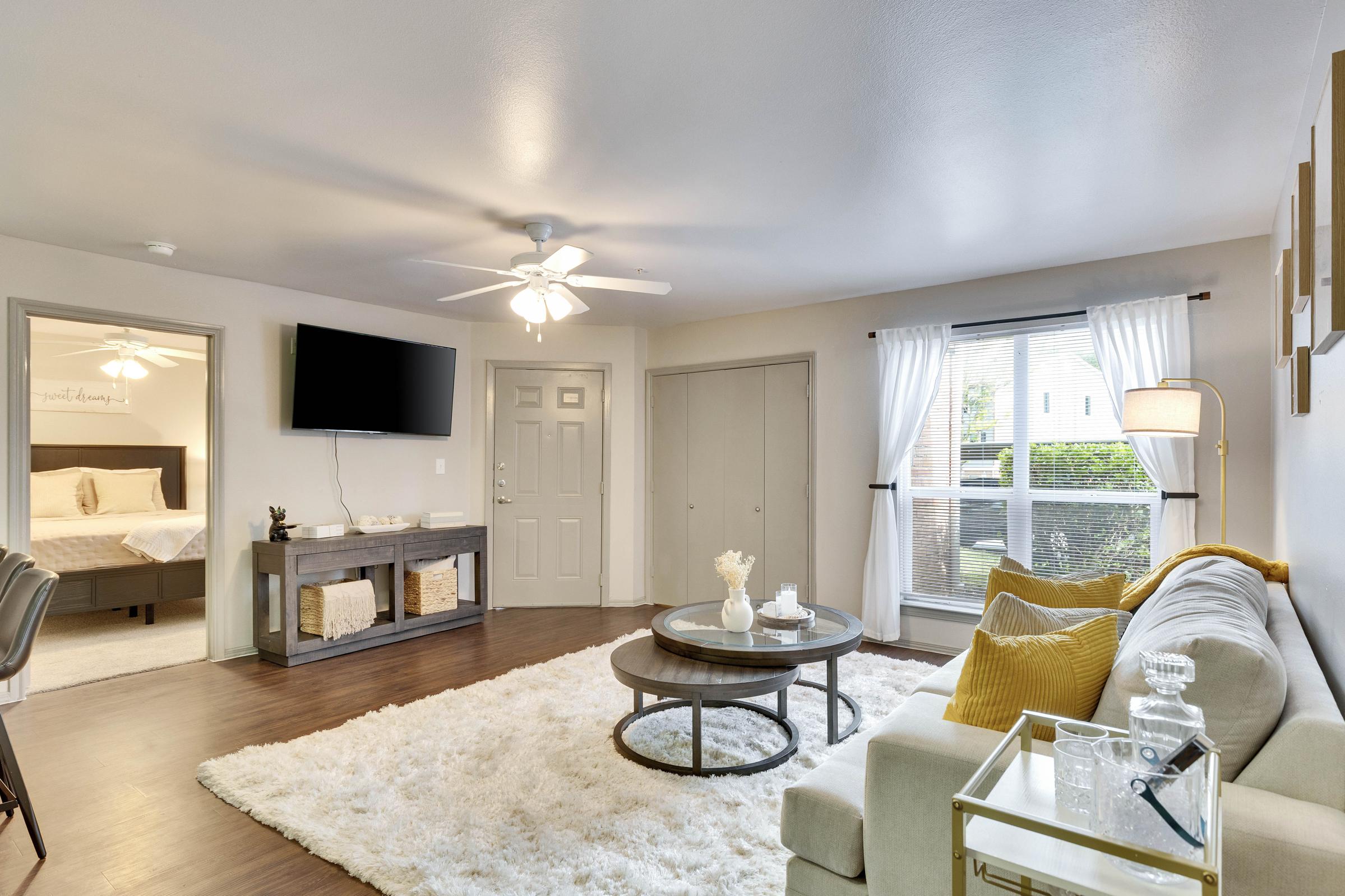 A cozy living room featuring a light-colored sofa with yellow pillows, a round coffee table on a plush white rug, and a mounted TV on the wall. The space is brightened by natural light from a window with sheer curtains and includes a dresser and an adjoining bedroom visible through an open door.