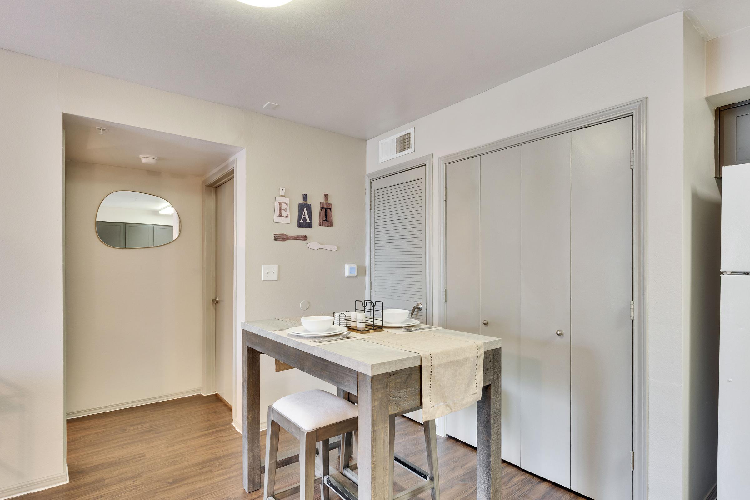 A modern kitchen area featuring a small wooden dining table with a light-colored tablecloth. Two white stools are positioned at the table. In the background, there’s a hallway with an oval mirror on the wall and two closed doors, along with decorative wall art that spells "EAT." The floor is made of wood.