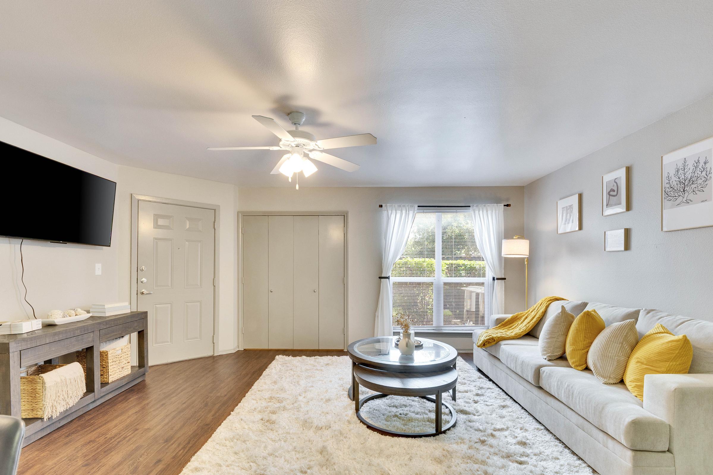 A cozy living room with a light gray sofa adorned with yellow pillows. There's a circular coffee table in front, and a large window with sheer curtains lets in natural light. A television is mounted on the wall, and a decorative console with storage baskets is visible. The floor is covered with a plush white rug.