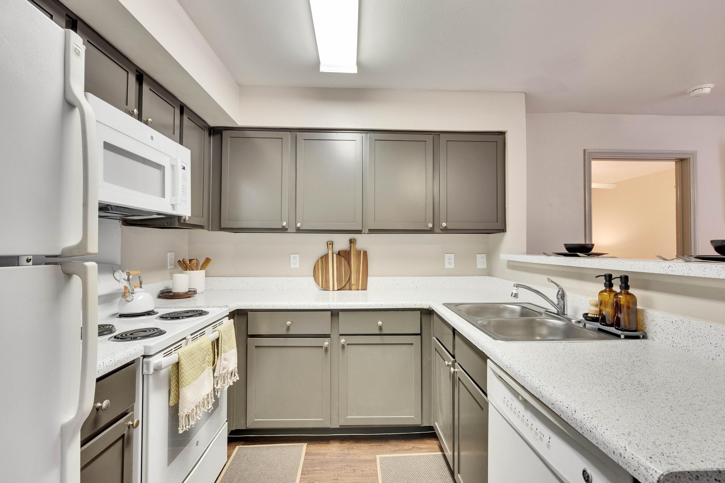 Modern kitchen featuring gray cabinetry, white countertops, and stainless steel appliances. Includes a microwave, stove, and sink with a wooden cutting board displayed. Minimalist decor with kitchen utensils and dishware visible, creating a clean and functional cooking space. Light-colored walls and a warm atmosphere.