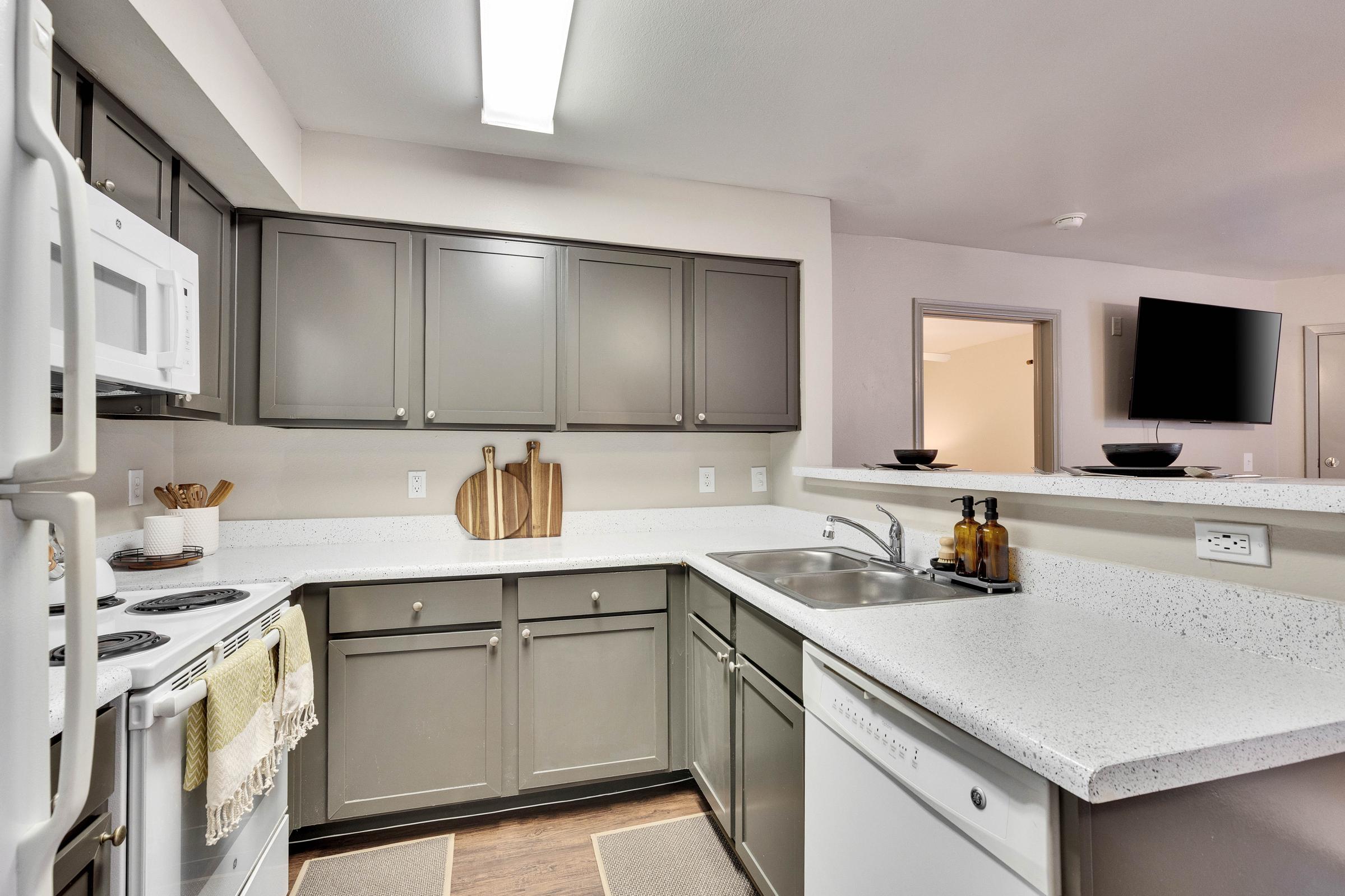 Modern kitchen with gray cabinets, a white countertop, and stainless steel appliances. Includes a microwave, stove, and sink with a cutting board and utensils displayed. A cozy atmosphere is created by a warm light and minimalist decor. The dining area is visible in the background with a television mounted on the wall.