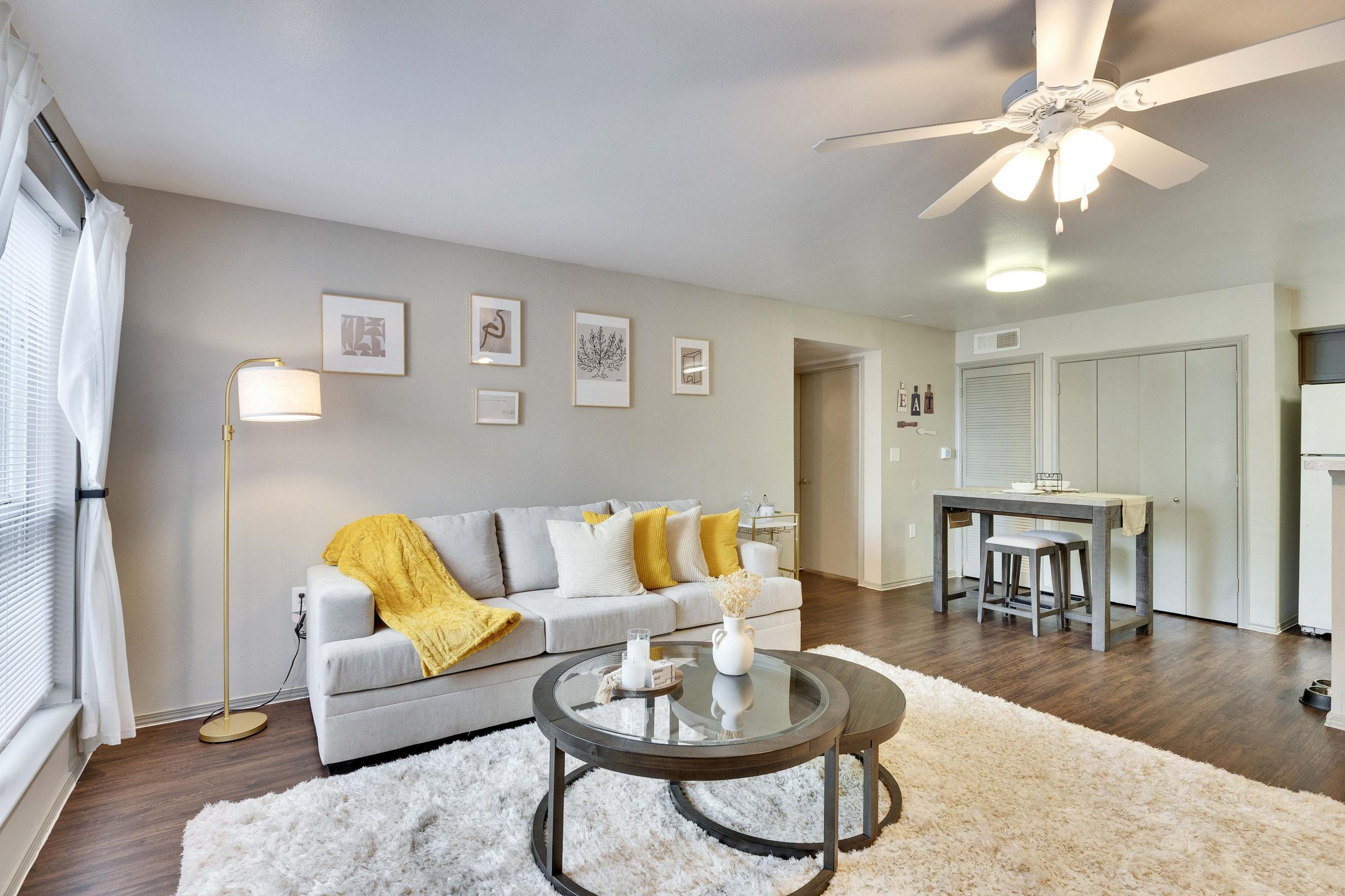 A cozy living room with a light gray couch adorned with yellow throw pillows and a blanket. A large, round coffee table sits on a soft, white rug. The walls are decorated with framed artwork, and a floor lamp stands next to the couch. A ceiling fan is visible, and there’s a dining table in the background.