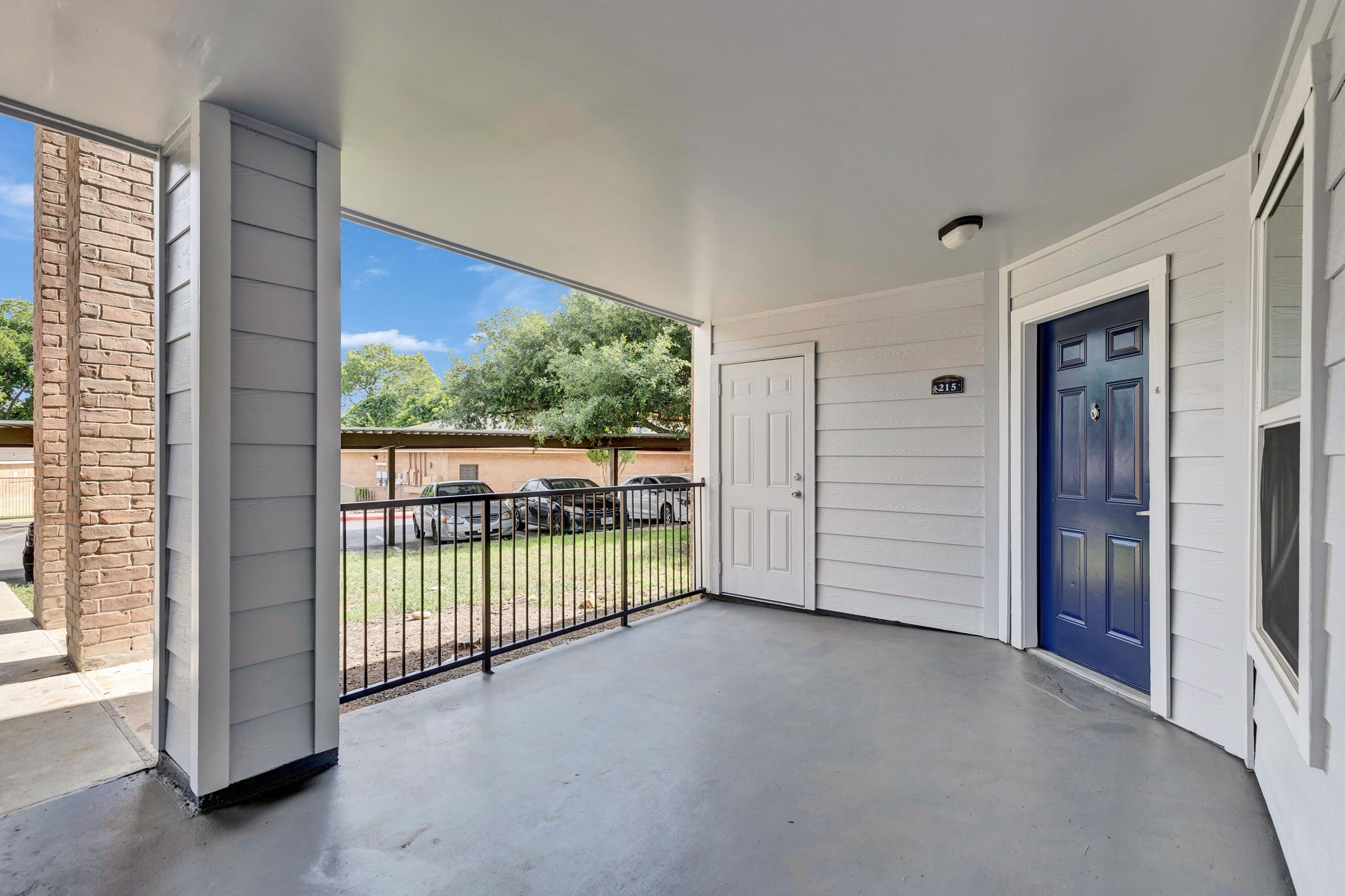 A covered porch area featuring a concrete floor, blue door on the right side, and a fence. Trees and a parking area are visible in the background with cars parked nearby. The space is well-lit, reflecting a sunny day with clear blue skies.