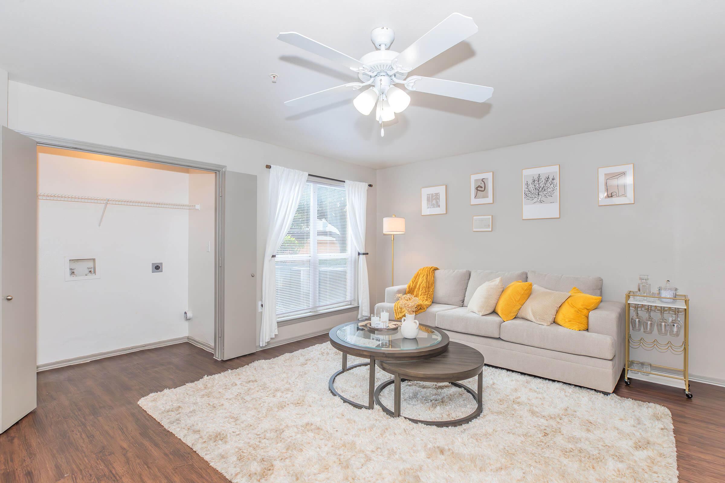 A cozy living room featuring a light gray sofa adorned with yellow pillows, a round coffee table with decor items, and a plush white area rug. Natural light streams in through a large window, complemented by sheer white curtains. A ceiling fan enhances the airy feel, and a closet is visible in the background.