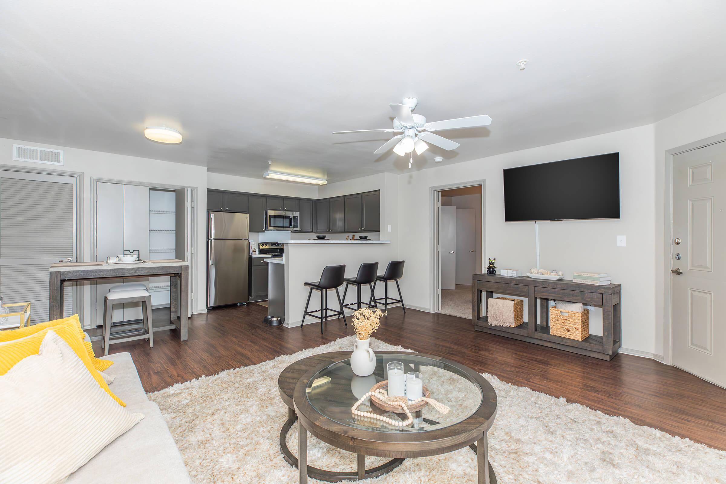 A modern, spacious living room featuring a cozy seating area with a beige sofa and a circular coffee table on a fluffy rug. The open kitchen is visible in the background, equipped with stainless steel appliances. A large flat-screen TV is mounted on the wall, and barstools are arranged near the kitchen counter.