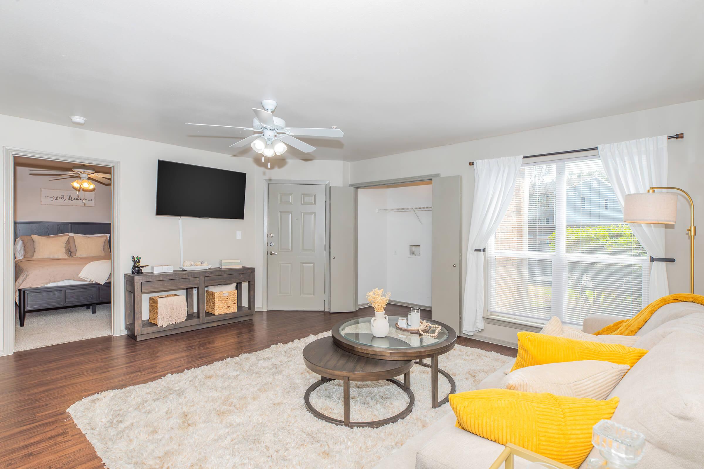 A cozy living room featuring a beige sofa with yellow accent pillows, a circular coffee table, and a plush area rug. A flat-screen TV is mounted on the wall, and a doorway leads to a bedroom. Large windows with white curtains allow natural light, enhancing the inviting atmosphere.