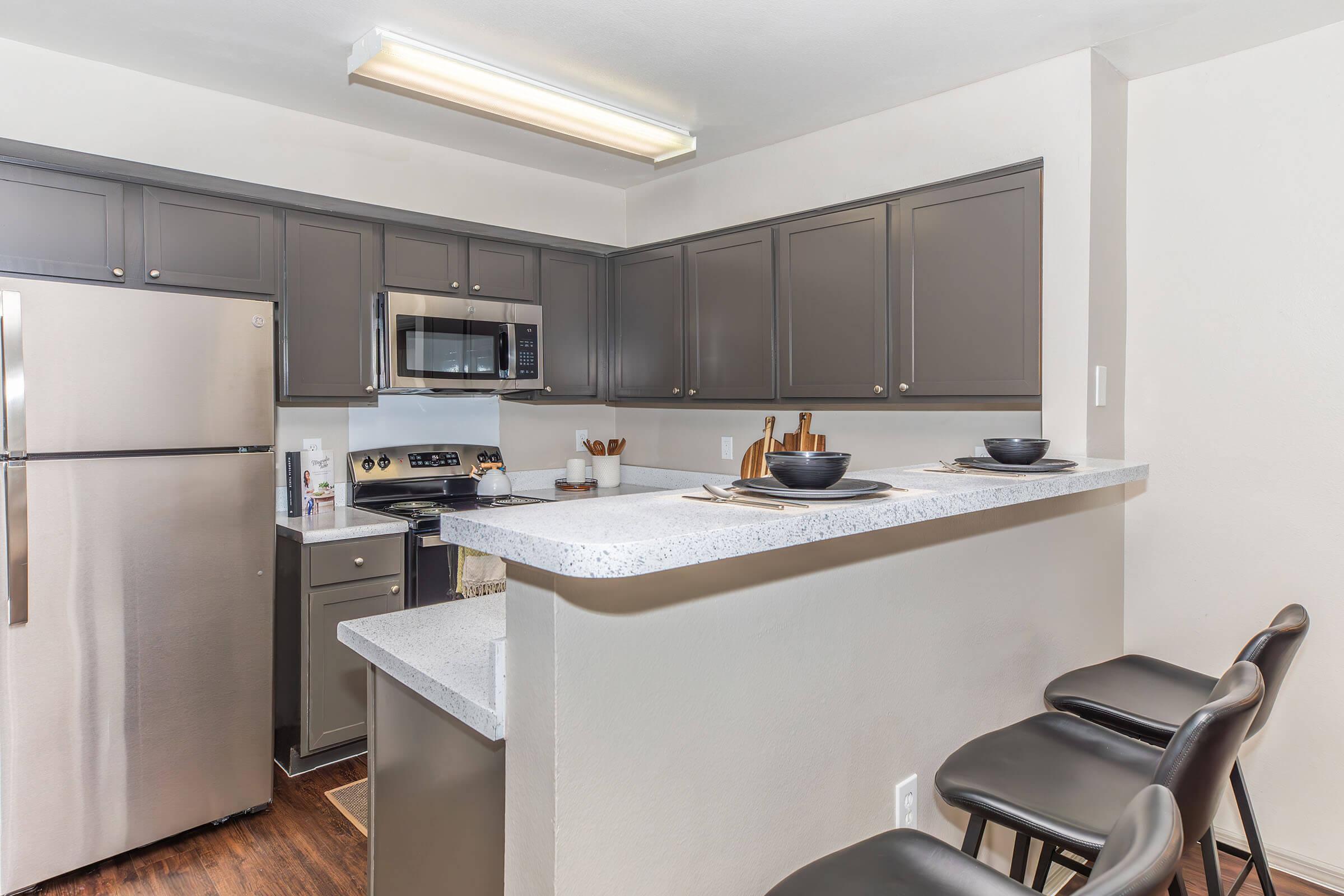 Modern kitchen featuring gray cabinets and stainless steel appliances. A large countertop separates the kitchen from the dining area, with three black bar stools positioned alongside. The space is well-lit with overhead lighting, creating a clean and inviting atmosphere.