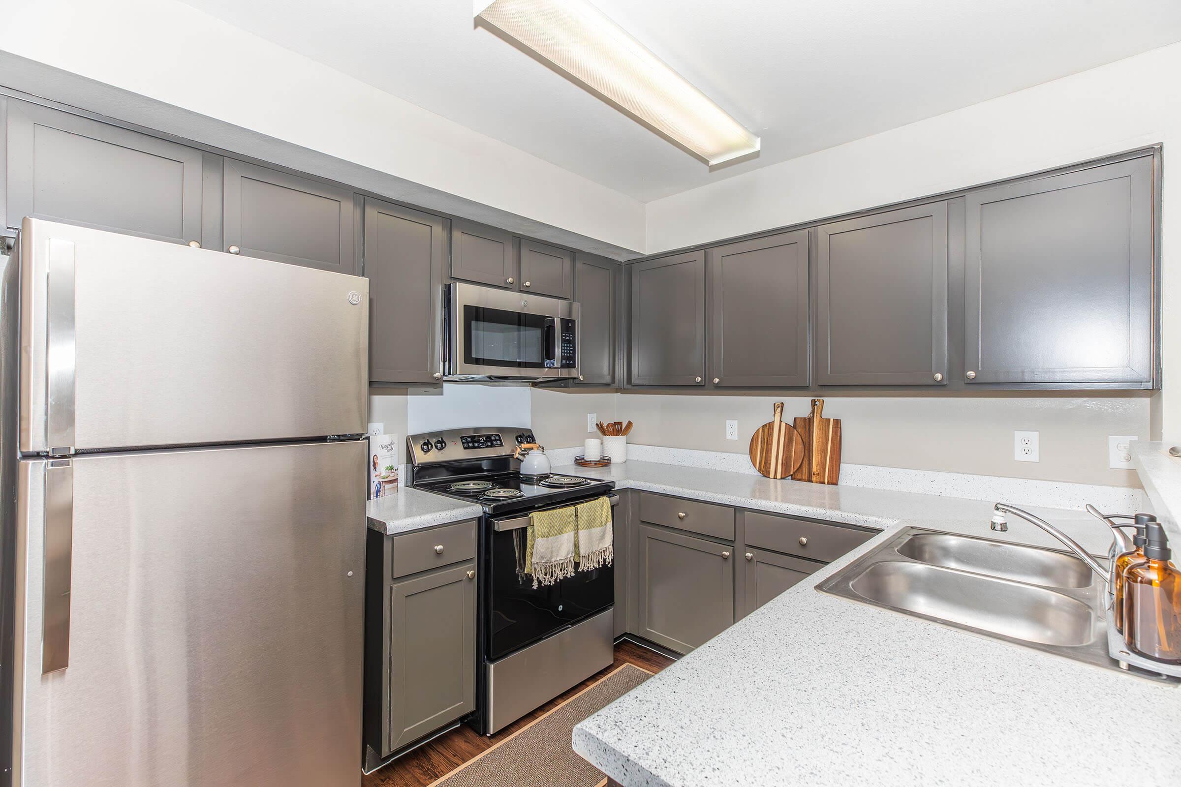 Modern kitchen featuring stainless steel appliances including a refrigerator and oven. The cabinets are gray, and the countertops are light-colored with a textured finish. A sink is visible beside the stove, and there are wooden cutting boards and kitchen utensils on the countertop. Natural light illuminates the space.