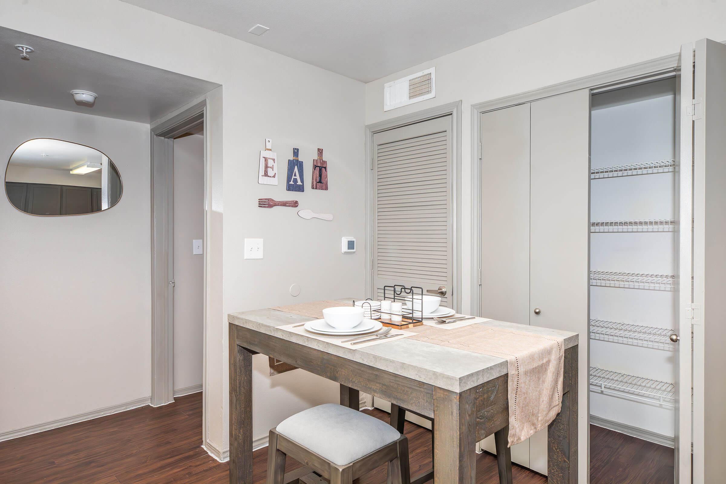 A cozy dining area featuring a small wooden table set for two, with plates and cutlery. The wall displays decorative letters spelling "EAT" alongside a wooden fork and knife. A mirror hangs nearby, and a closet door is partially open, revealing shelving inside. The overall decor is modern and minimalist.